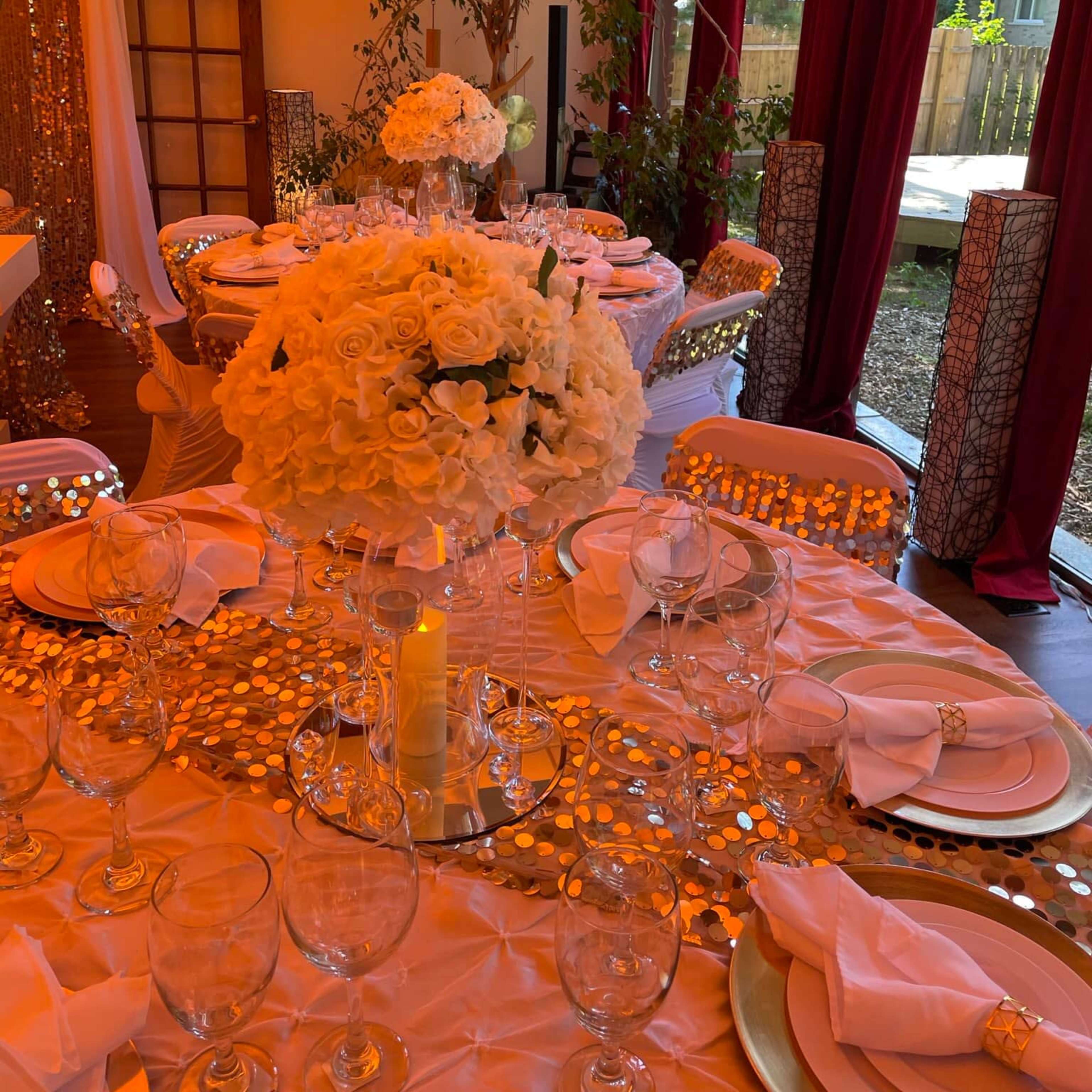 A dining table is elegantly set with white floral centerpieces, crystal glassware, and golden table decor under warm lighting.