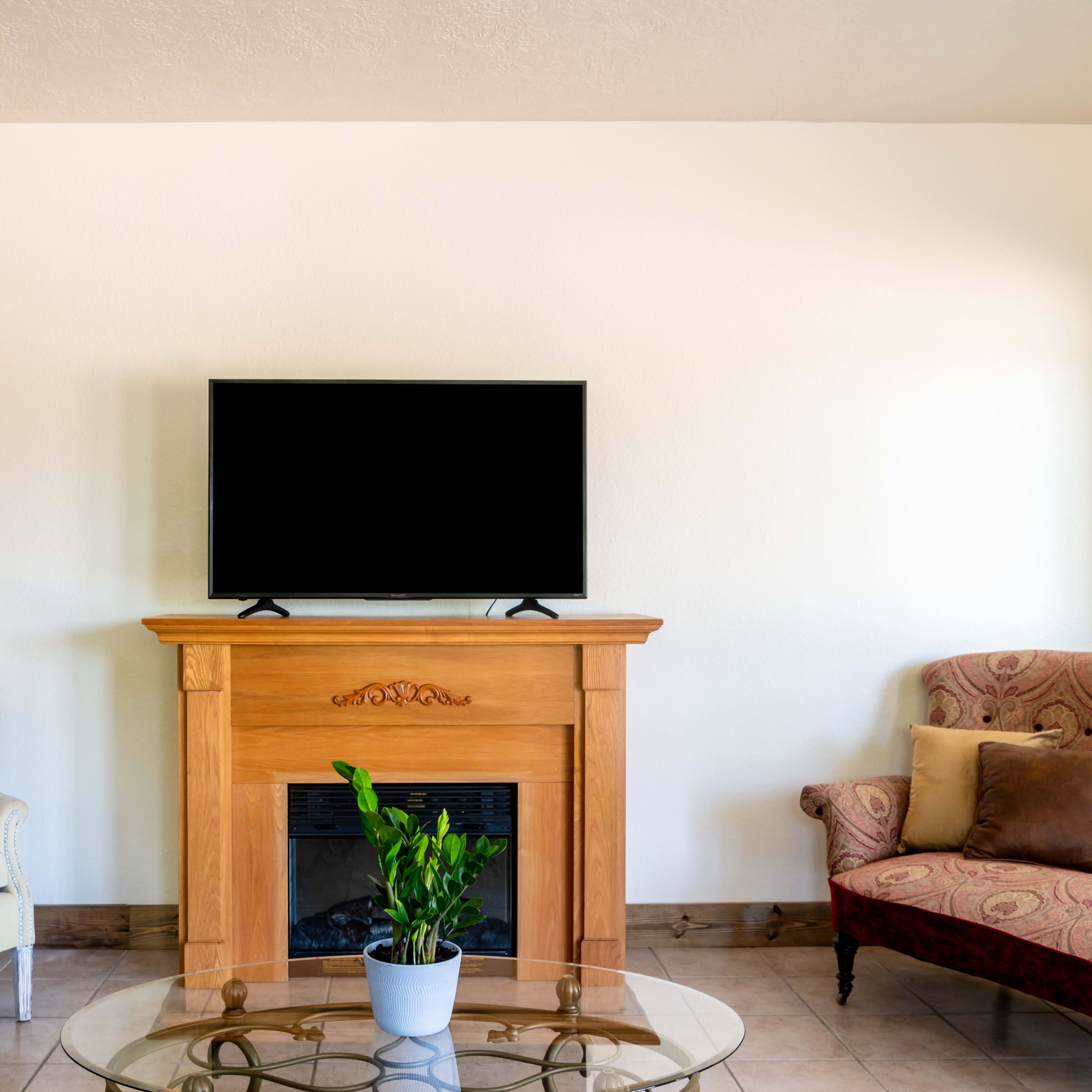 A living room with a wooden fireplace, a television mounted above it, and two chairs arranged around a glass coffee table.