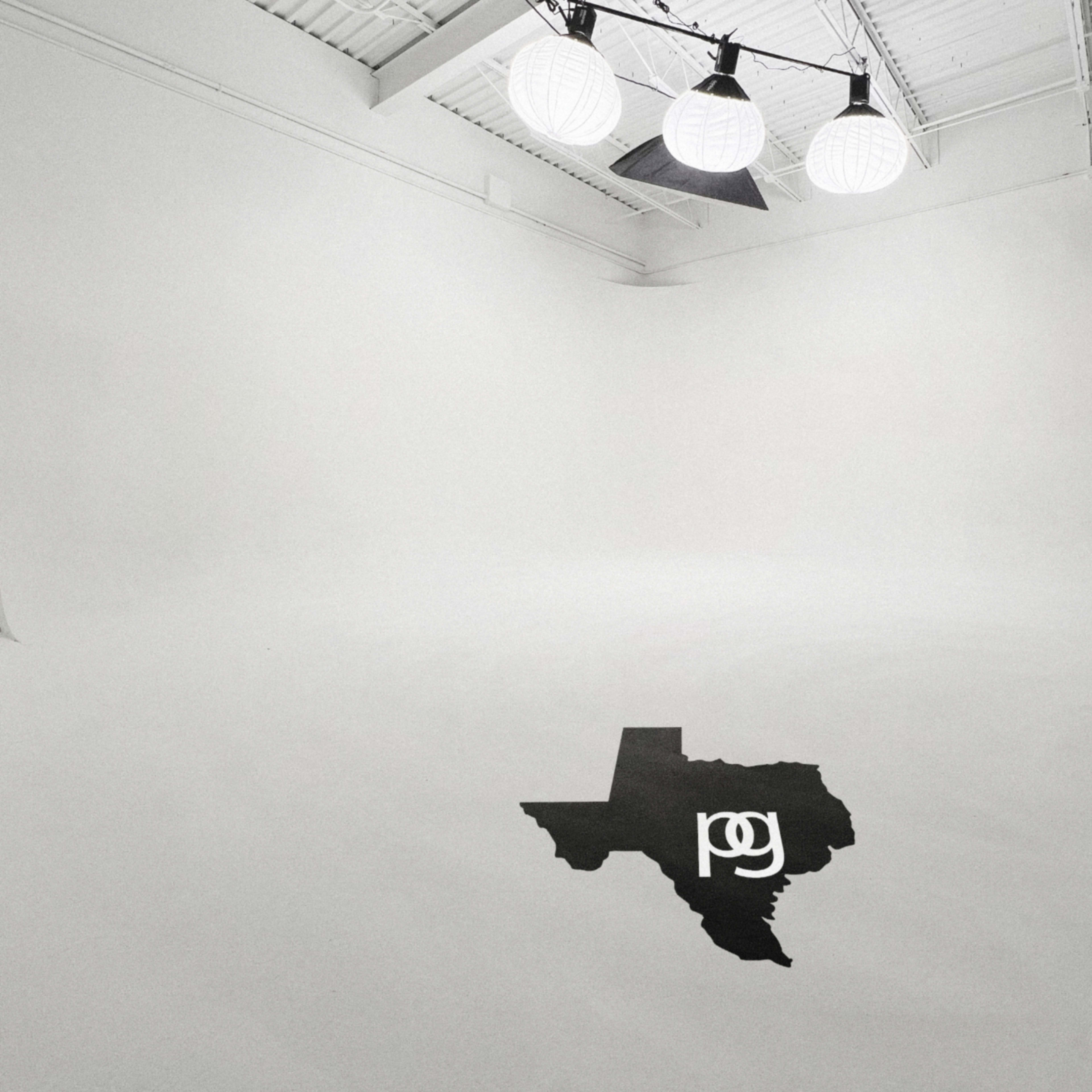 A spacious, empty studio with white walls and ceiling features three large round lights hanging above a black outline of the state of Texas on the floor.