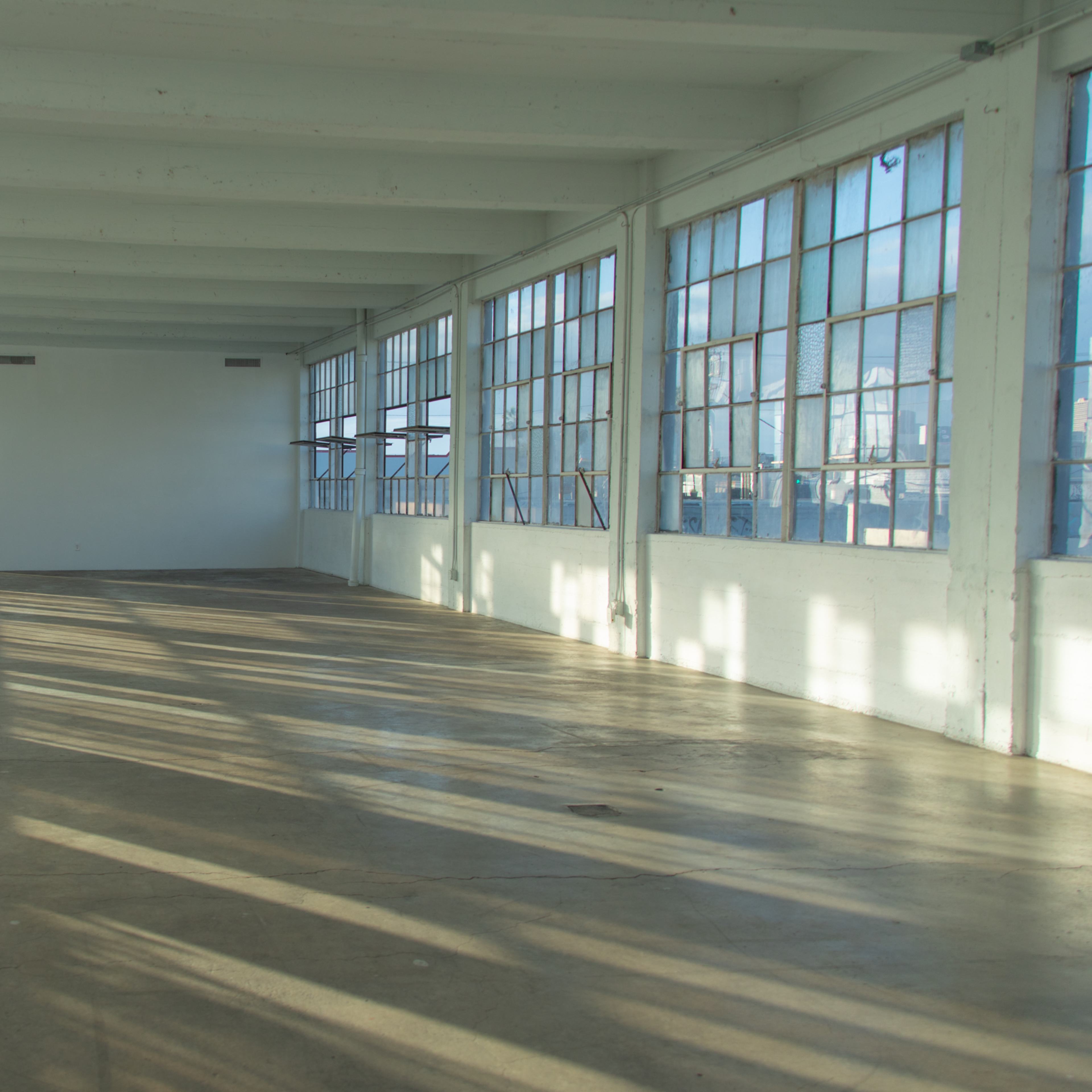 A spacious, empty room with large windows allowing natural light to fill the space, and concrete flooring.