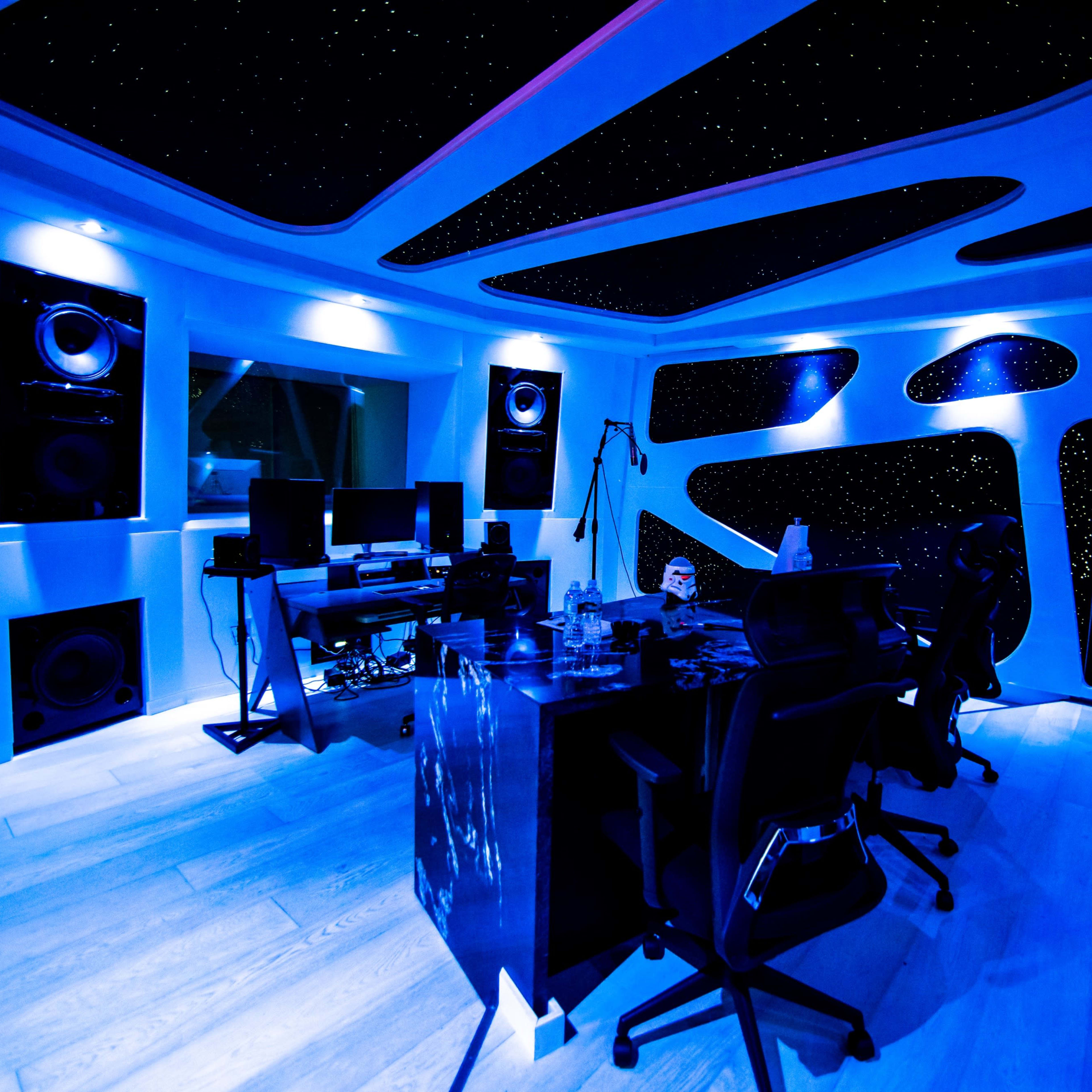 A music studio with a futuristic design, featuring dark blue lighting, large speakers, and equipment arranged around a central desk.