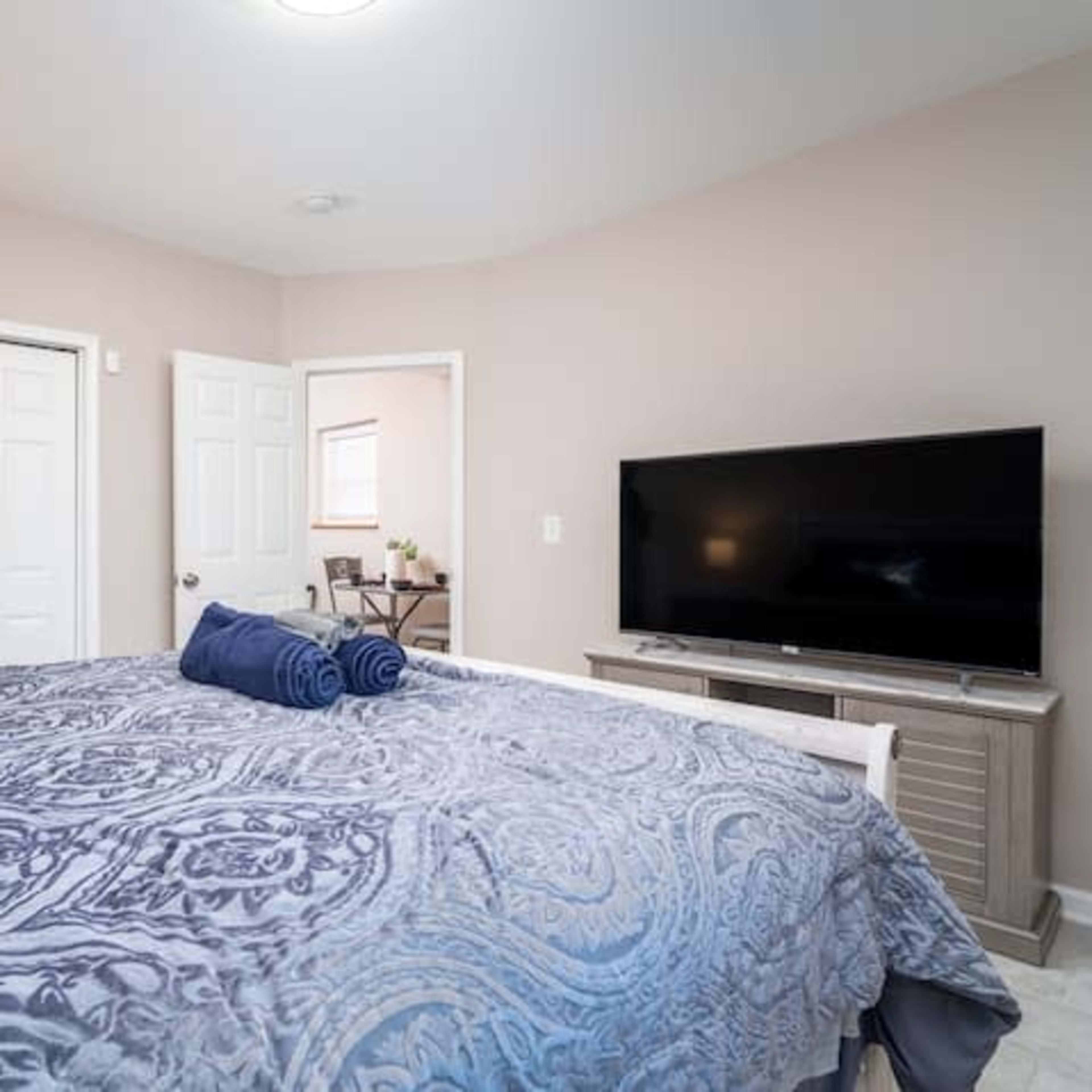 A spacious bedroom features a large bed with a patterned comforter, neatly rolled towels, a television on a stand, and a doorway leading to another room.