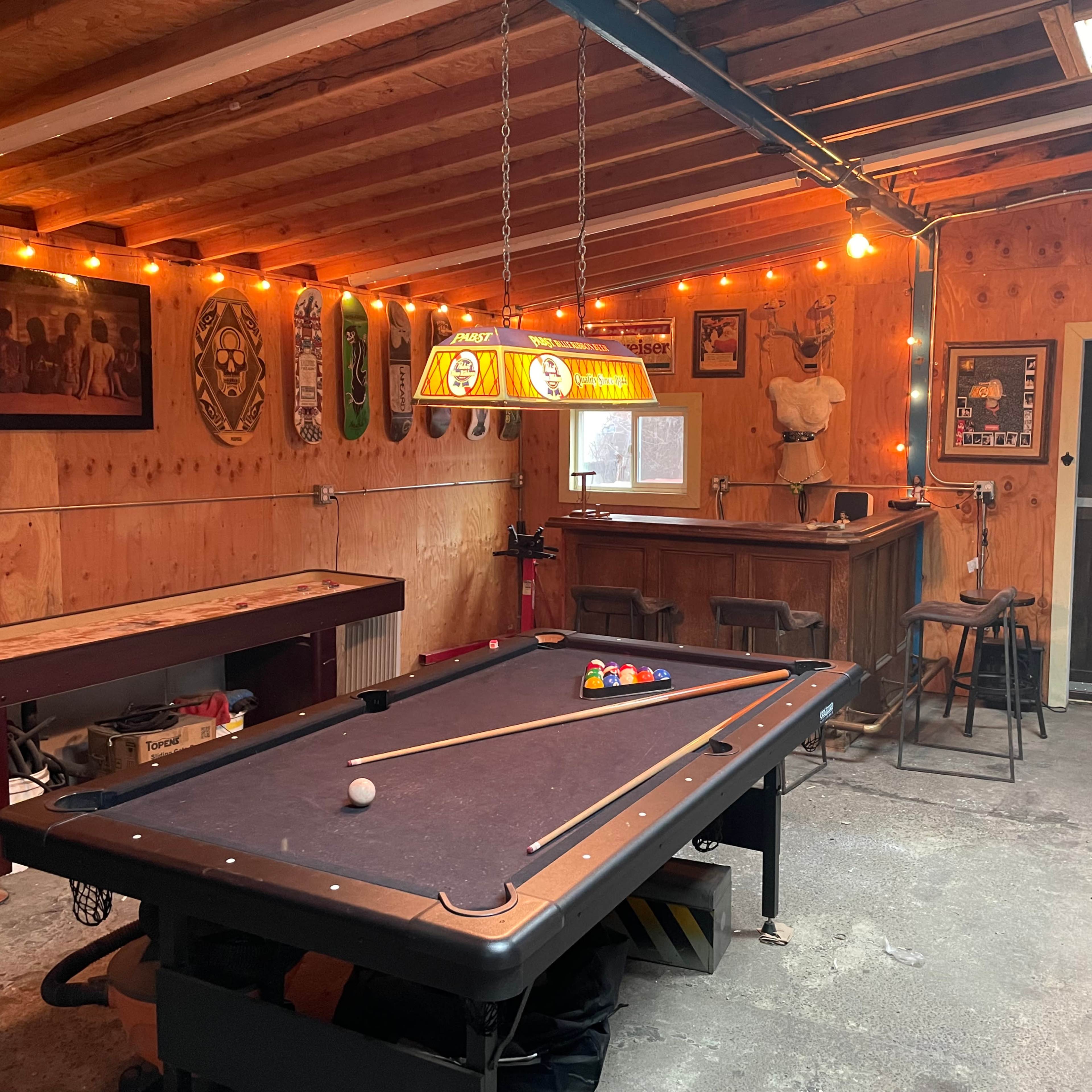 A rustic game room with a pool table, bar area, and wooden walls illuminated by string lights.