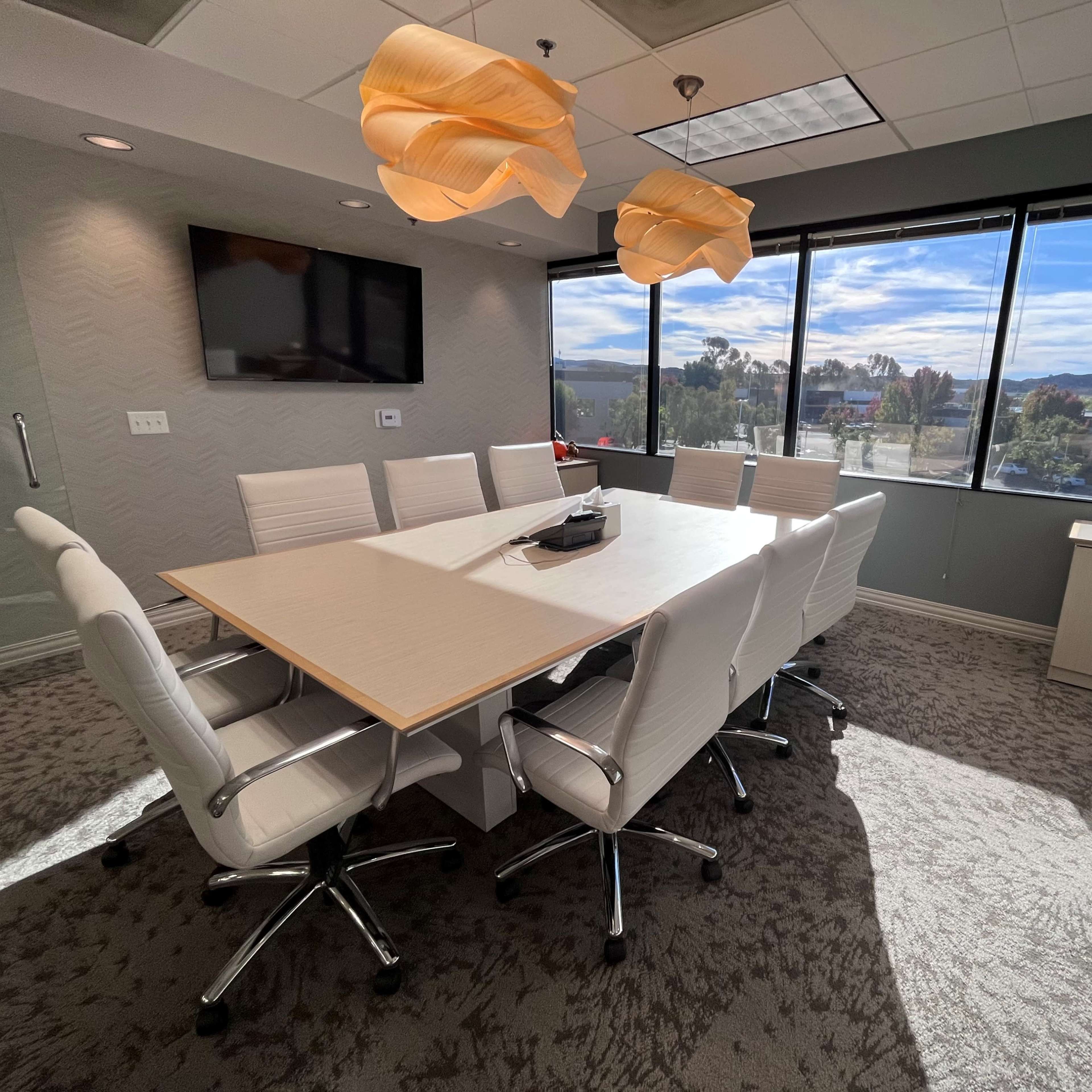 A modern conference room features a large table surrounded by white chairs, with windows offering a view of the outdoors and contemporary lighting fixtures above.