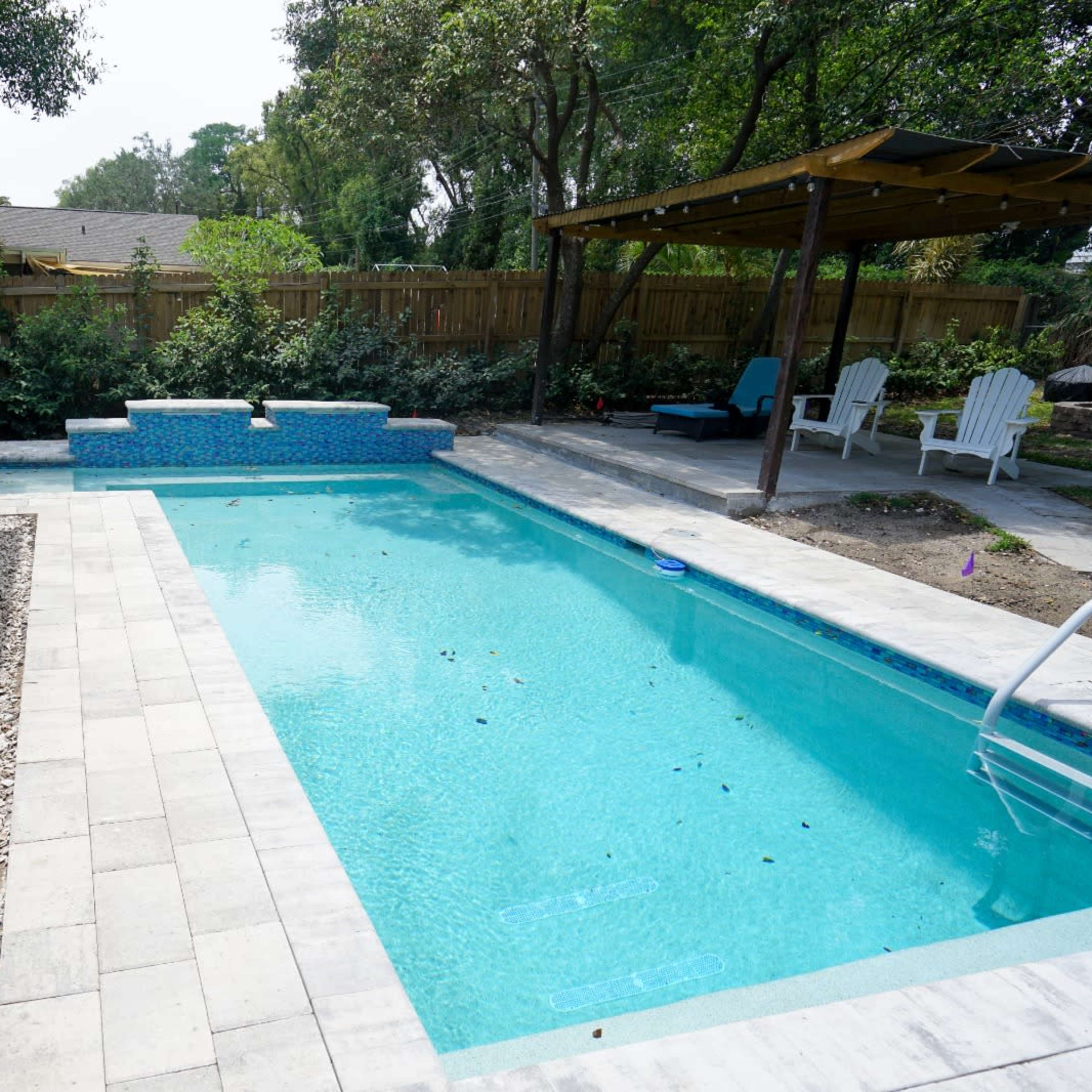 A backyard pool surrounded by patio tiles and a small seating area with white chairs under a shaded structure.
