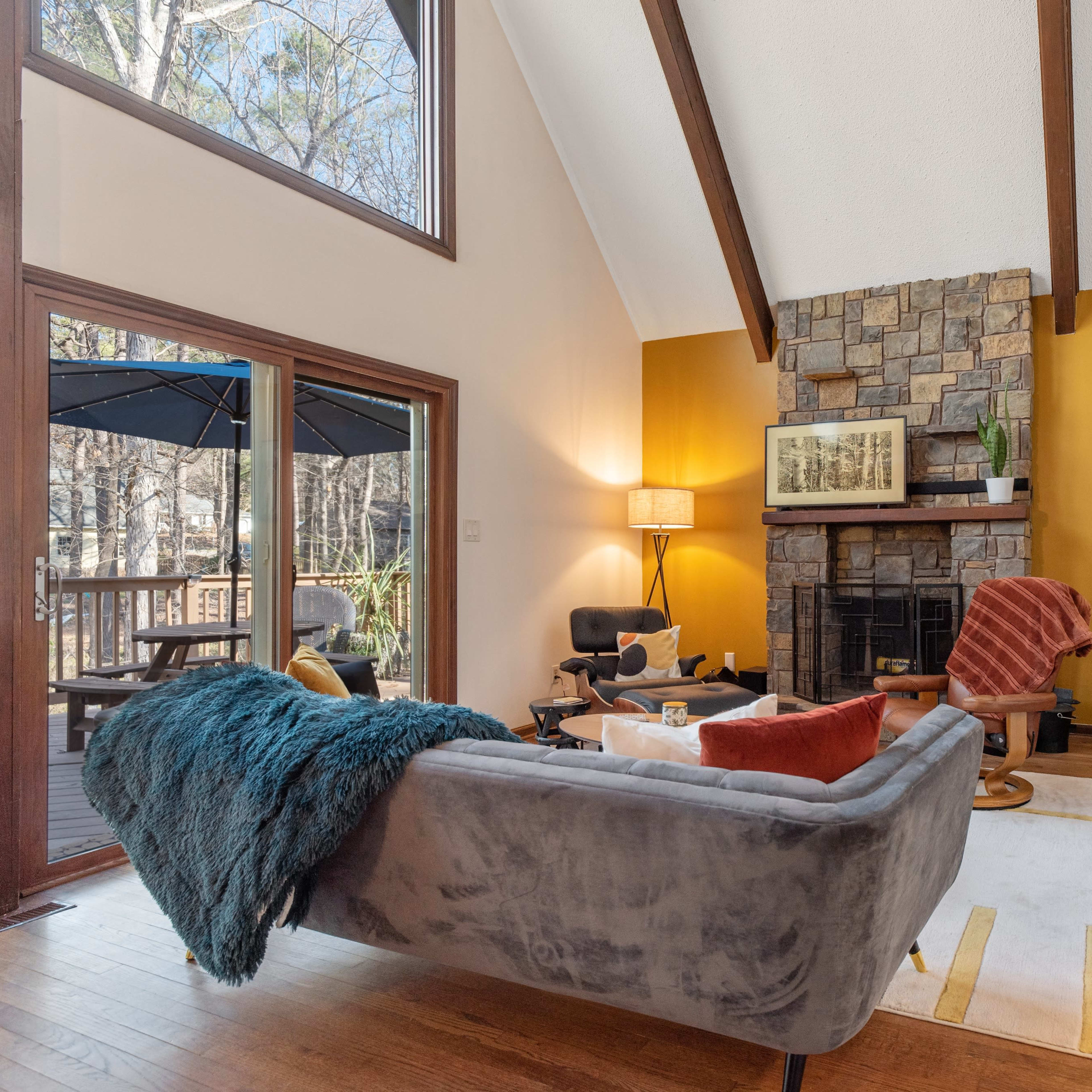 The living room features a gray sofa facing a stone fireplace, with a wooden staircase leading to an upper level and large windows allowing natural light to enter.