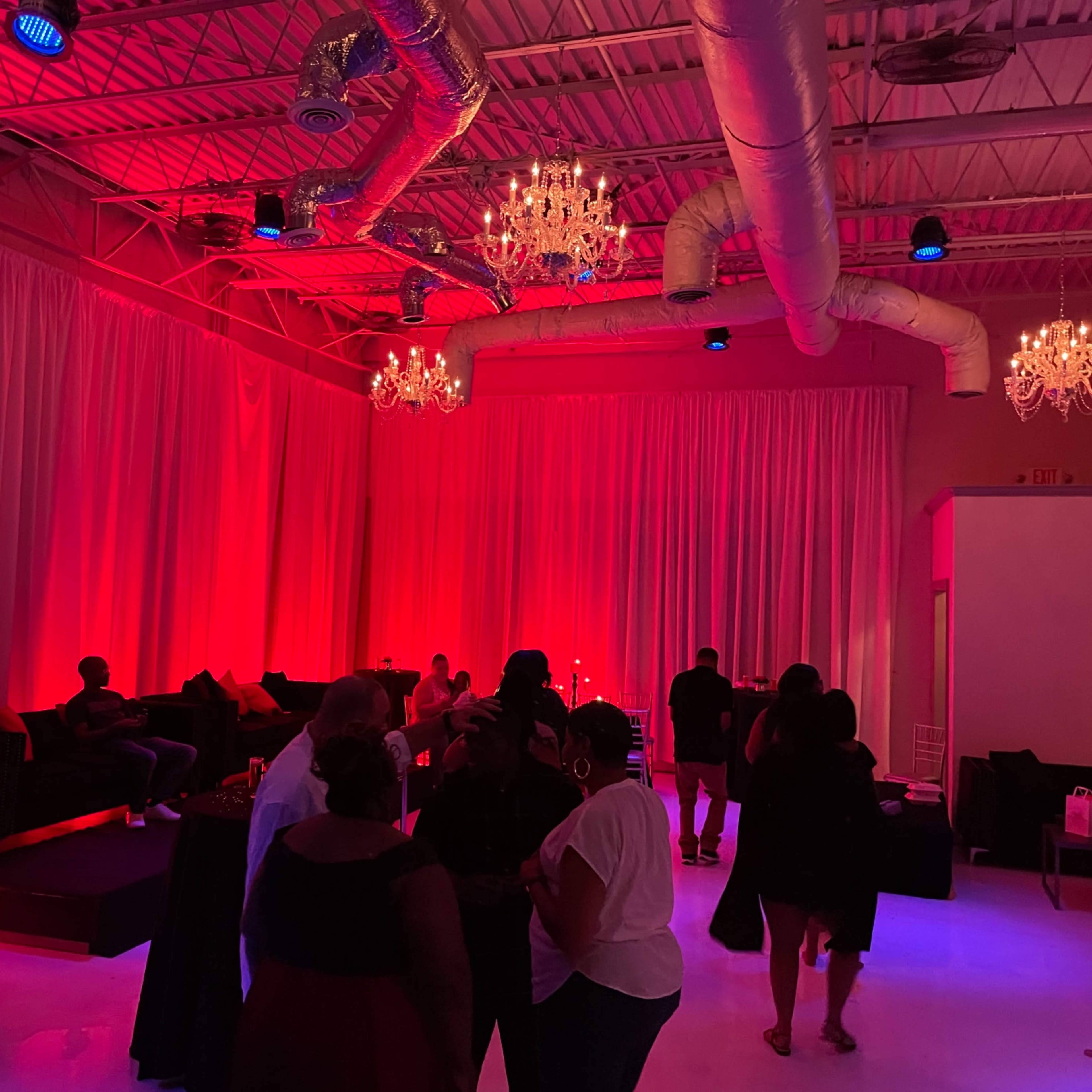 A dimly lit event space features red lighting, chandeliers, and guests mingling among couches and tables.
