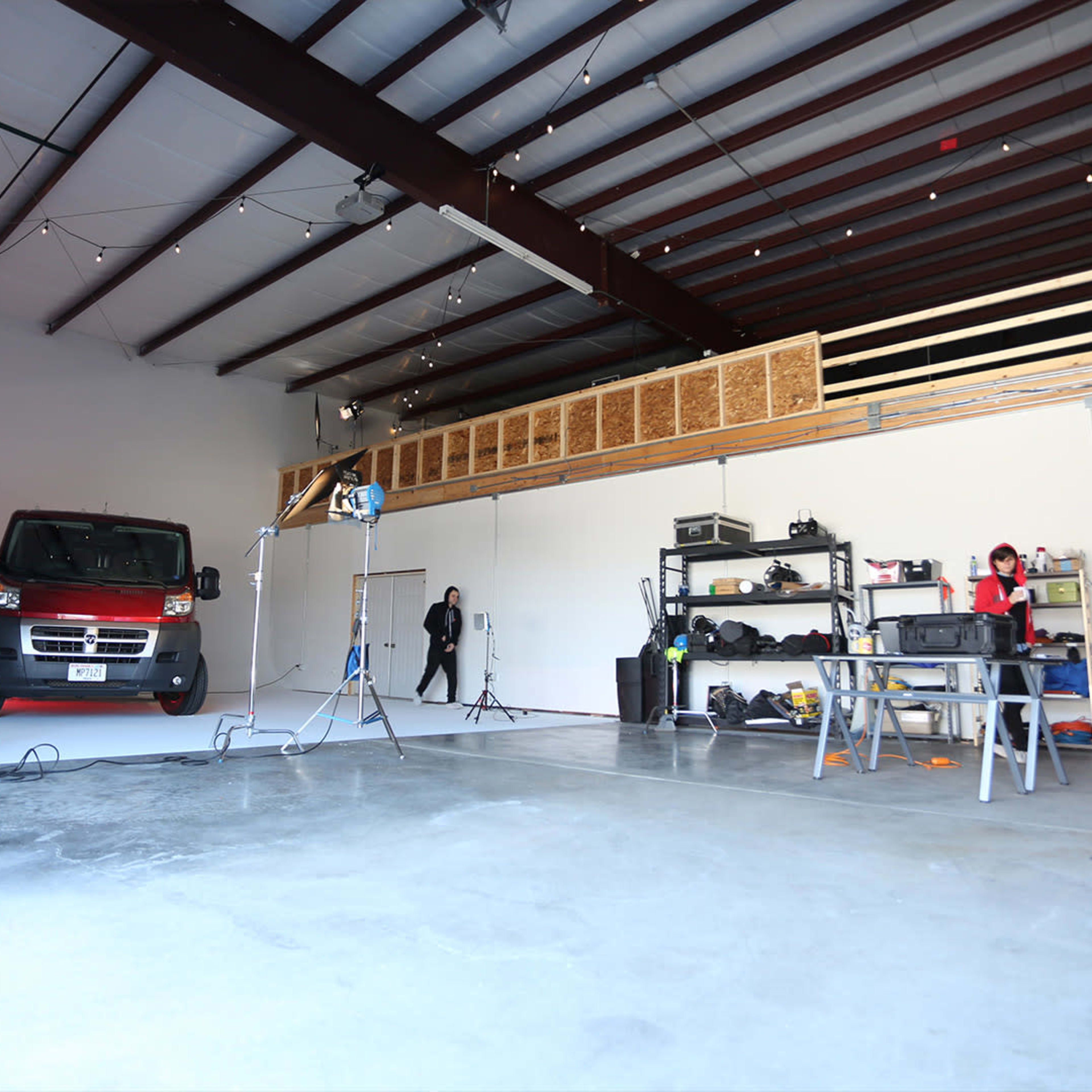 A spacious, well-lit warehouse with a red van and various lighting equipment set up for a production, accompanied by a work table and storage shelves.