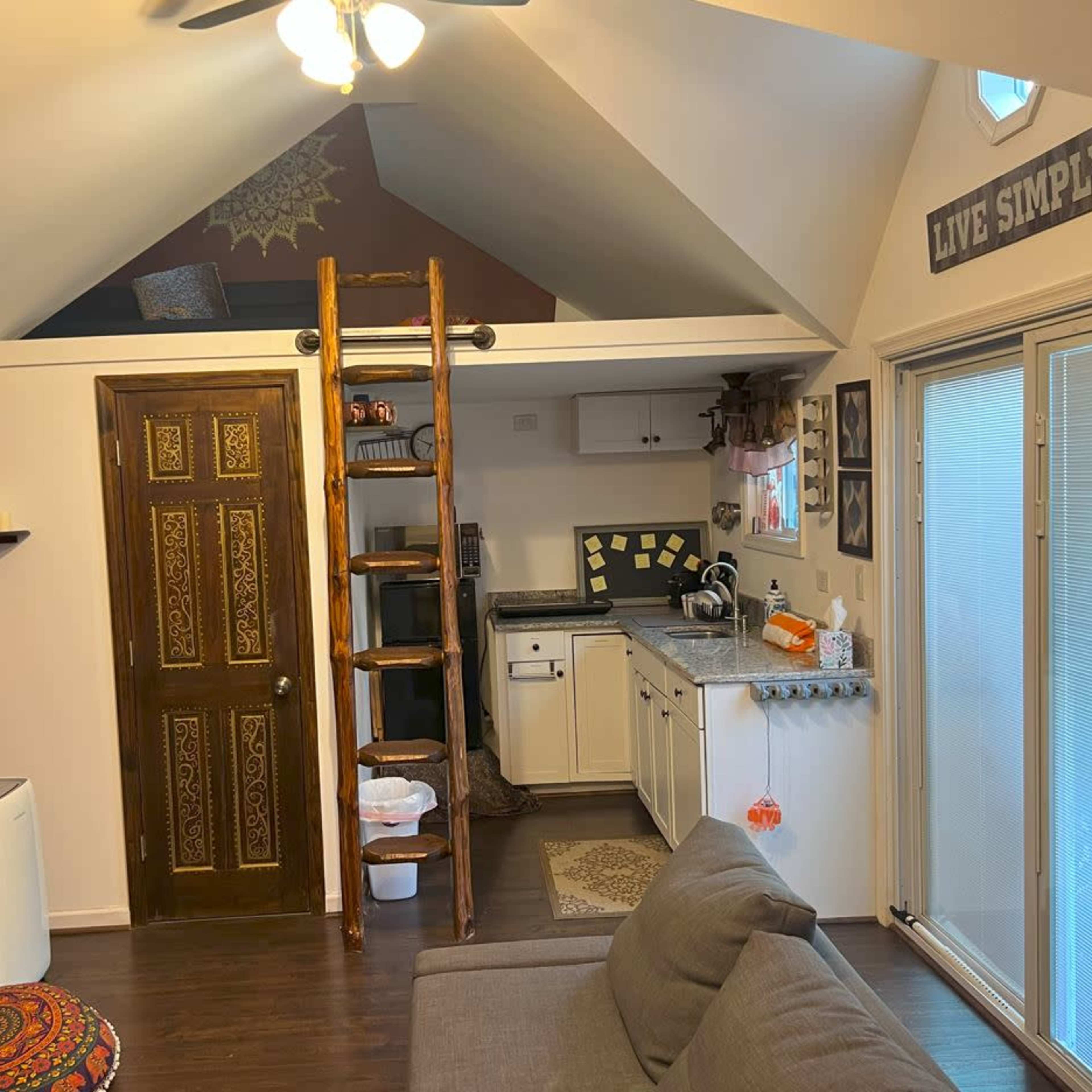 A small, cozy living area with a lofted sleeping space accessed by a wooden ladder, a kitchenette, and sliding glass doors leading outside.