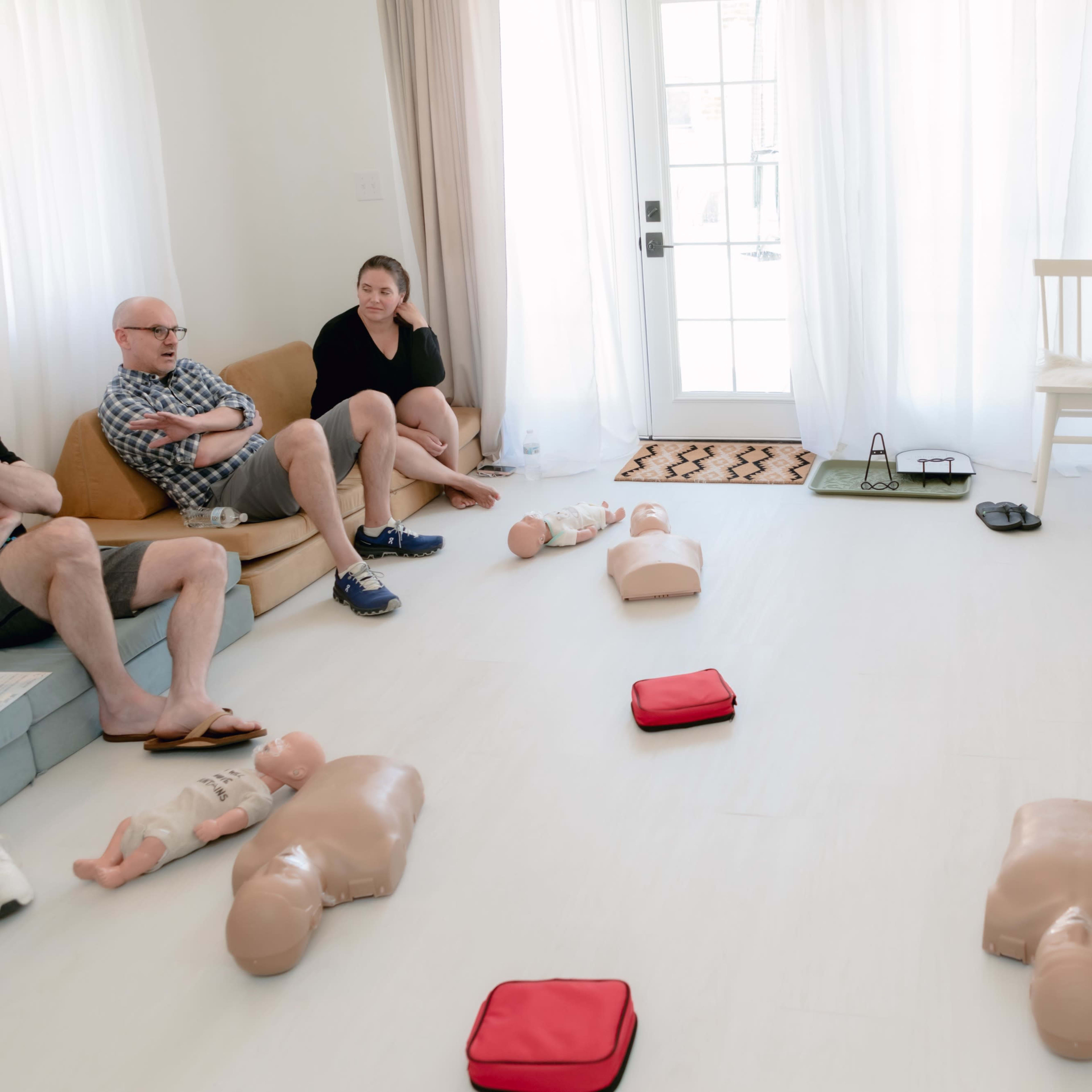A group of people is seated in a light-filled room with CPR training mannequins placed on the floor.