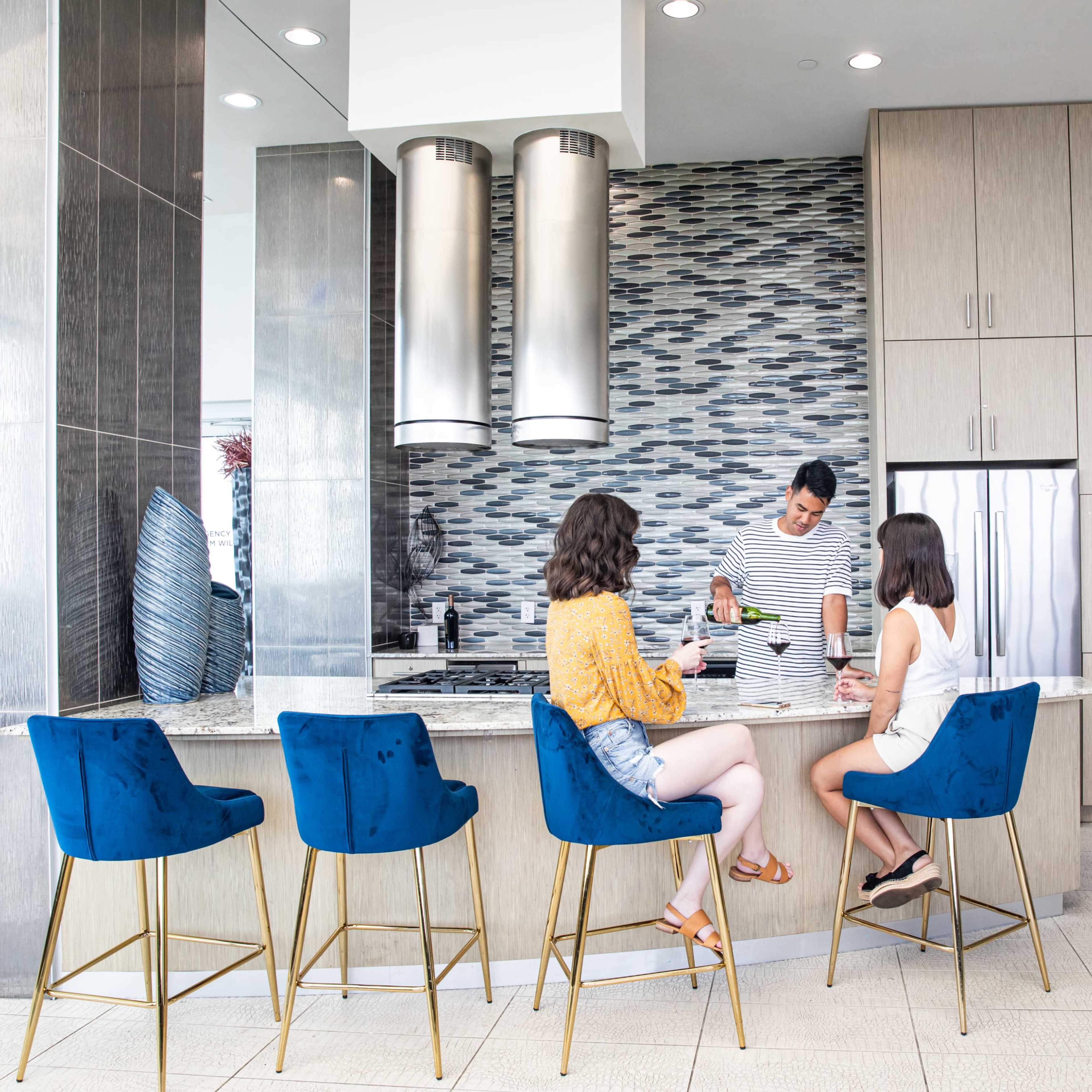 Two women sit at a modern bar counter while a male staff member prepares drinks in a stylish kitchen area.
