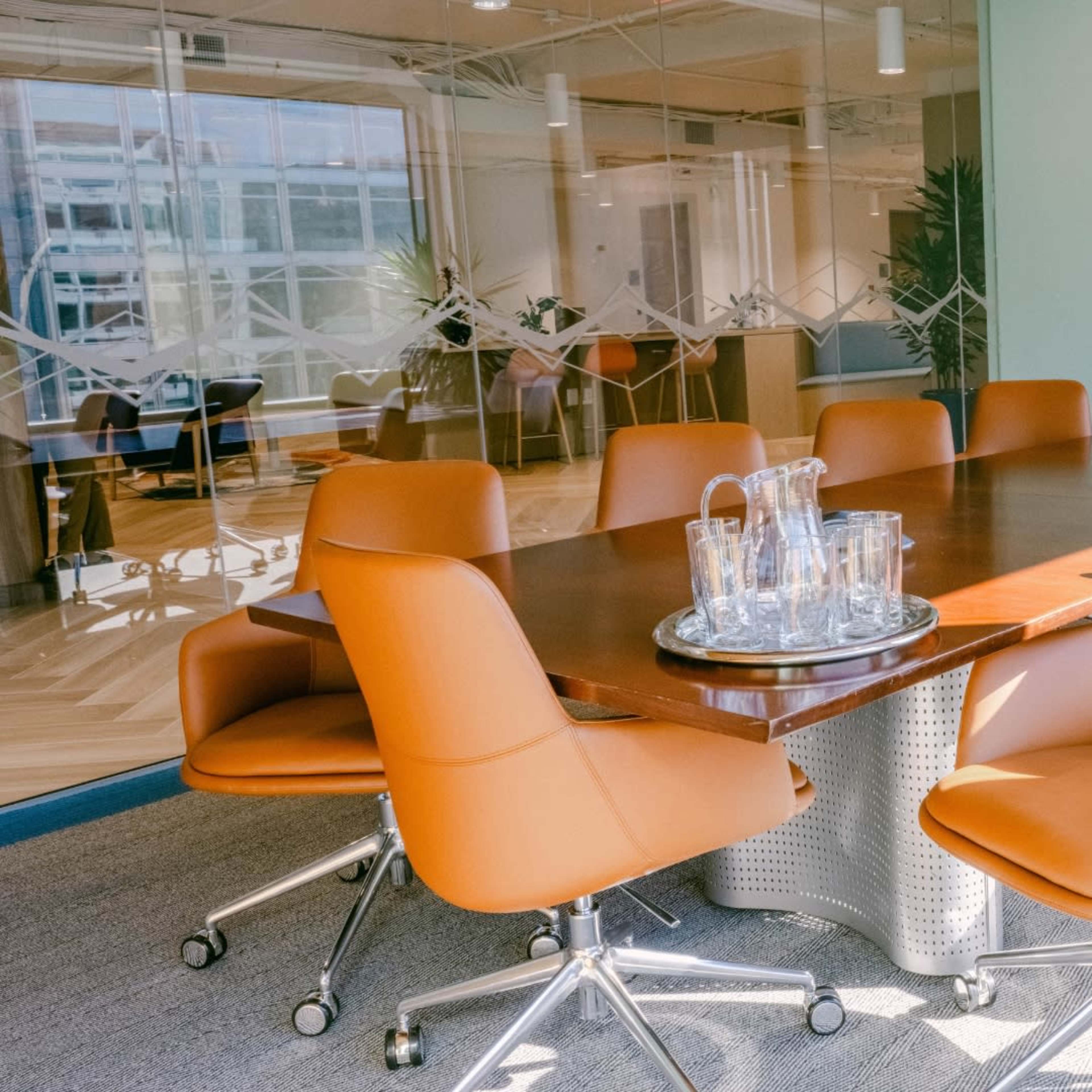 A modern conference room features a rectangular wooden table surrounded by eight orange leather chairs and glass water pitchers on a tray.