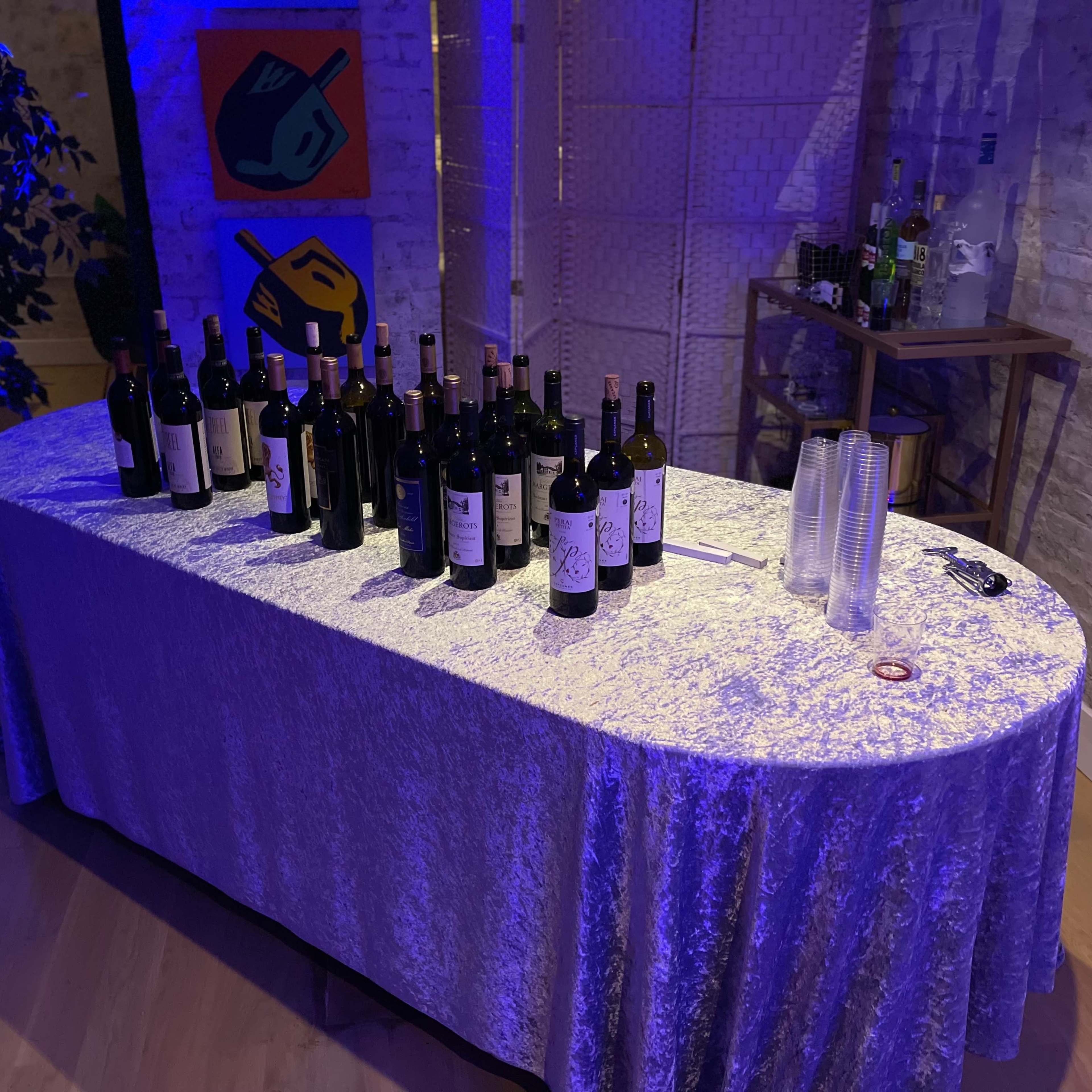 A table covered with a silver tablecloth displays numerous bottles of wine and plastic cups under blue lighting in a room with exposed brick walls.