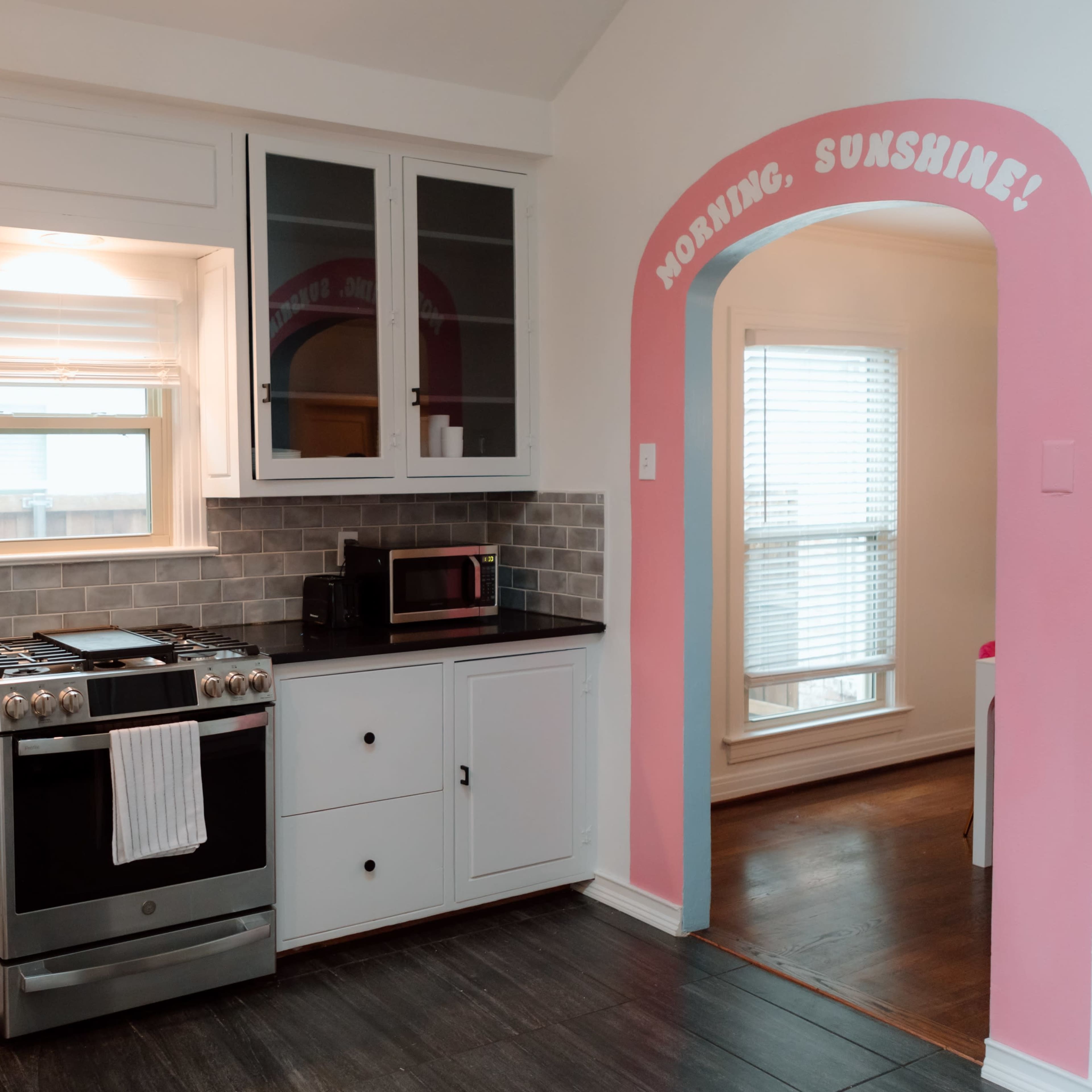 A modern kitchen with black countertops, white cabinets, a stainless steel stove, and an adjacent pink archway with the text "MORNING, SUNSHINE!" leading to another room.