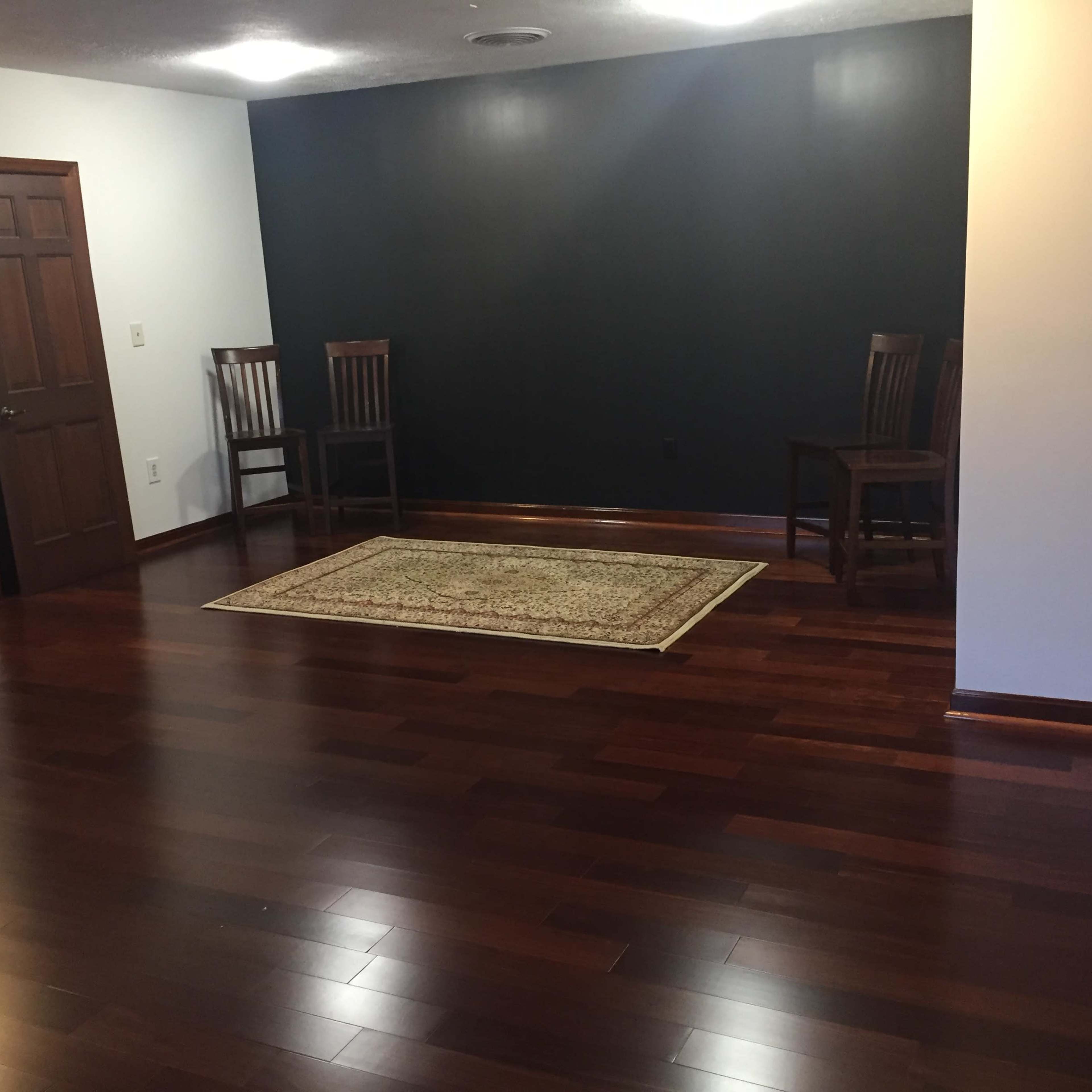 A room with dark wooden flooring, a patterned rug in the center, two wooden chairs on one side, and a dark feature wall.