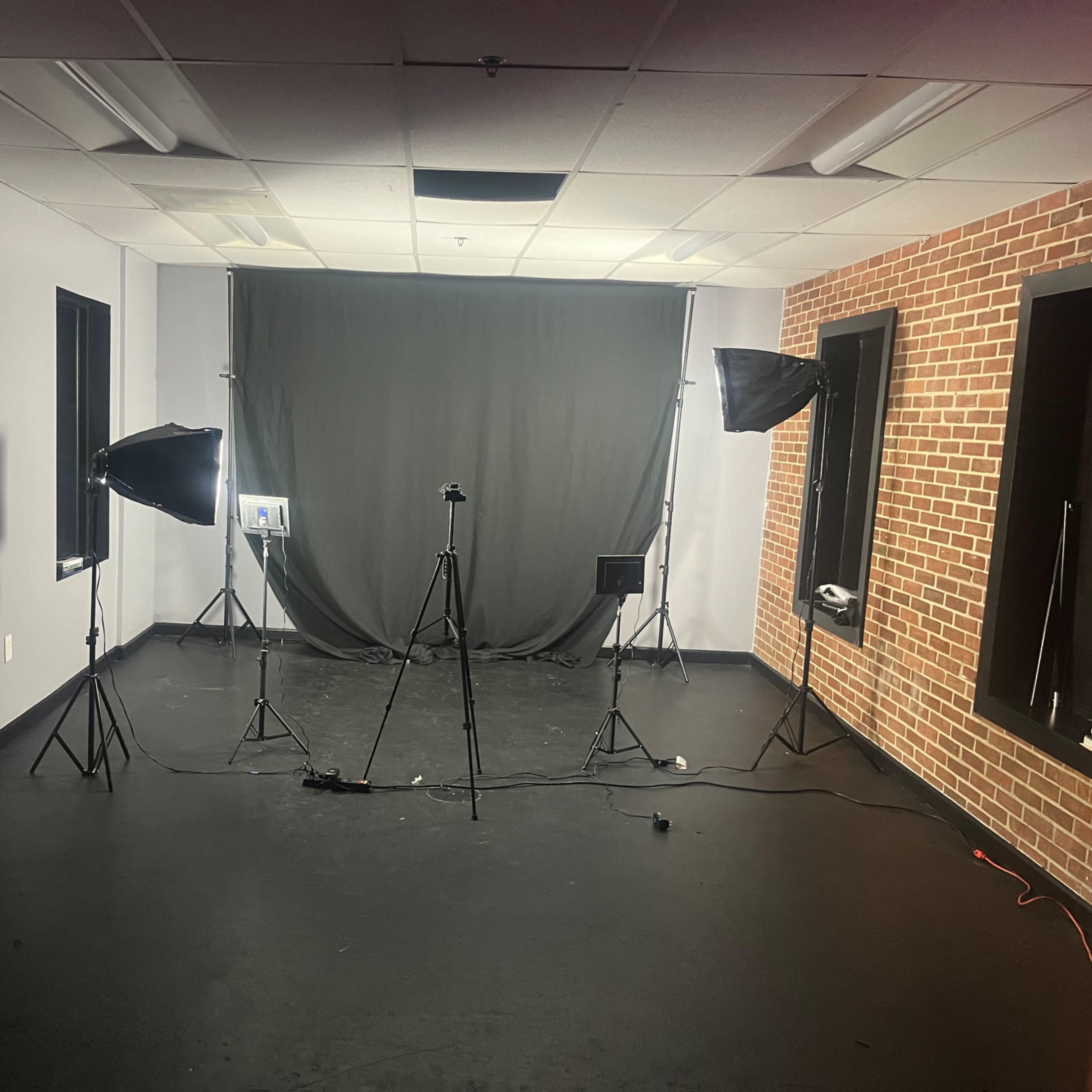 A dimly lit studio setup with a gray backdrop, two softbox lights on stands, and a camera tripod positioned in front.