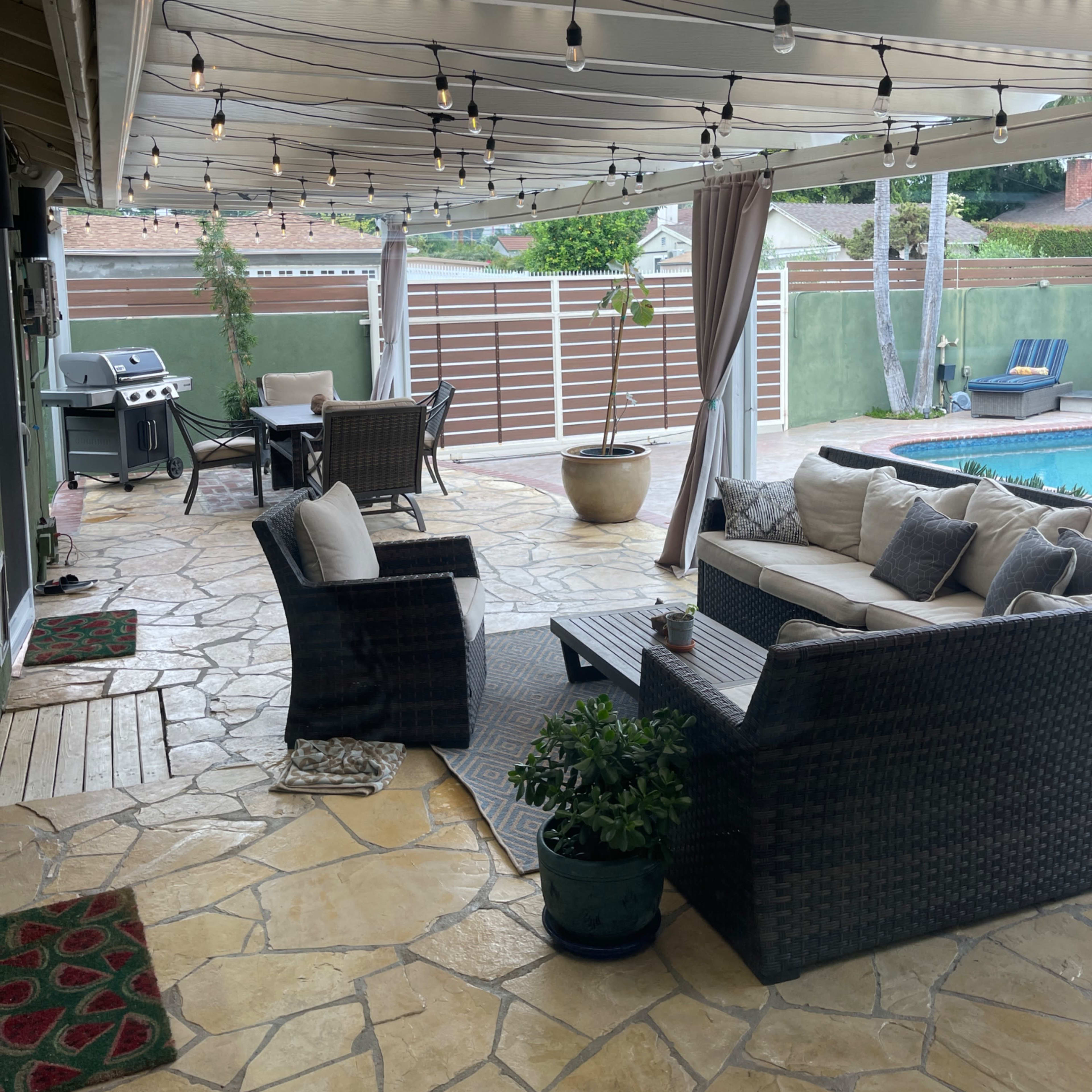 A patio area with a stone floor, outdoor furniture, a grill, and decorative string lights overhead, adjacent to a swimming pool.