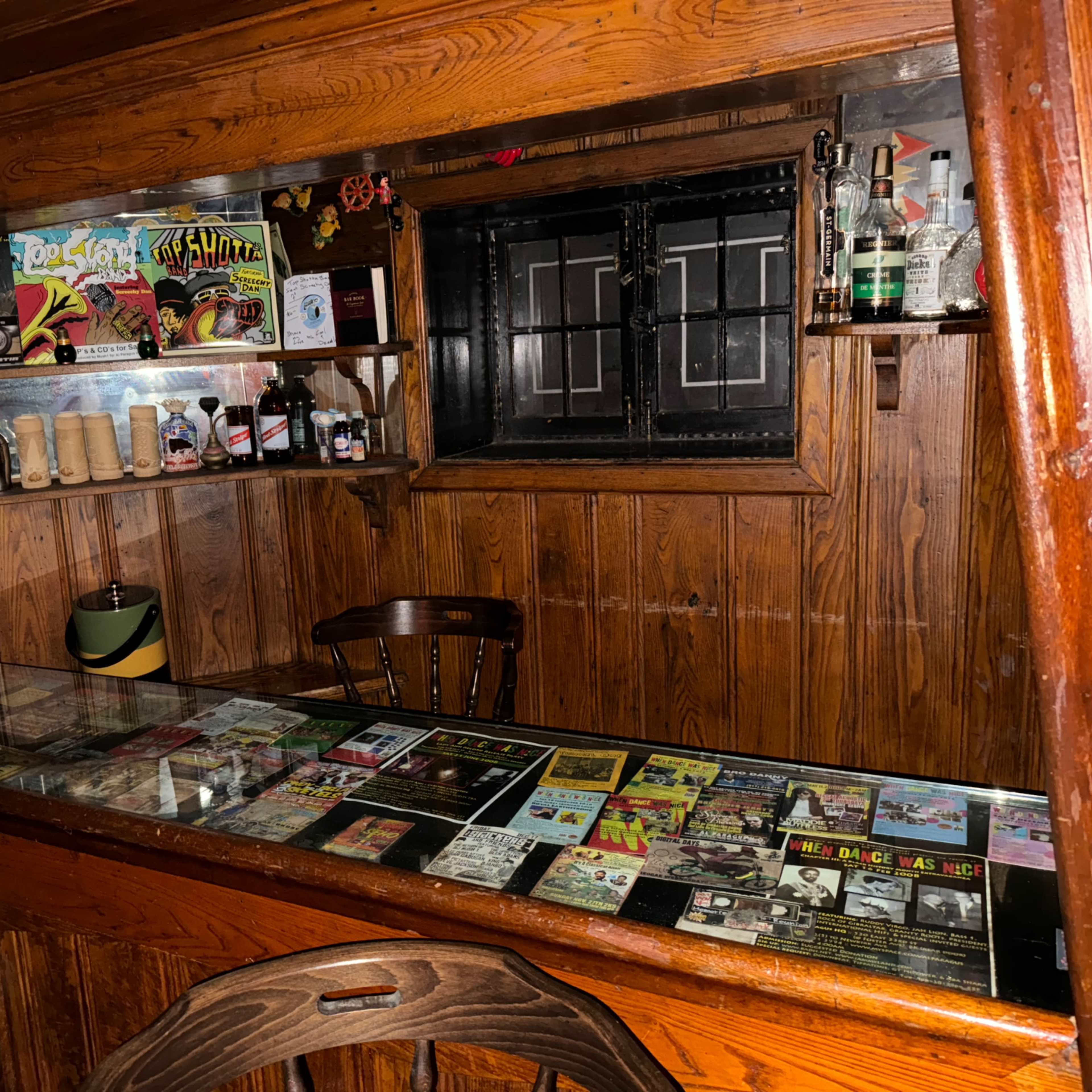 A wooden bar with a glass countertop displays vintage magazines and a shelf containing various bottles and glassware.
