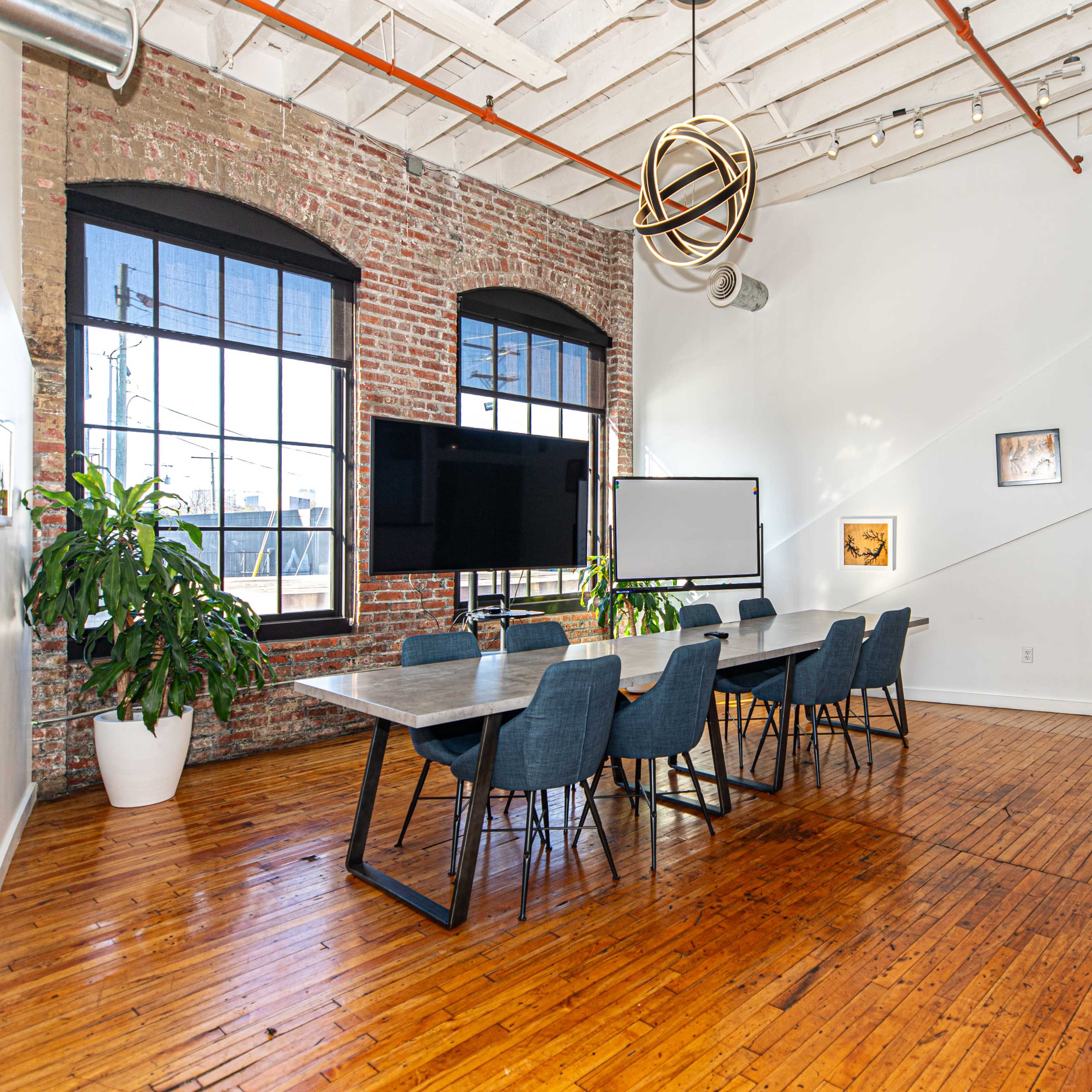A spacious conference room features a long table surrounded by chairs under industrial-style windows, with exposed brick walls and wooden floors.