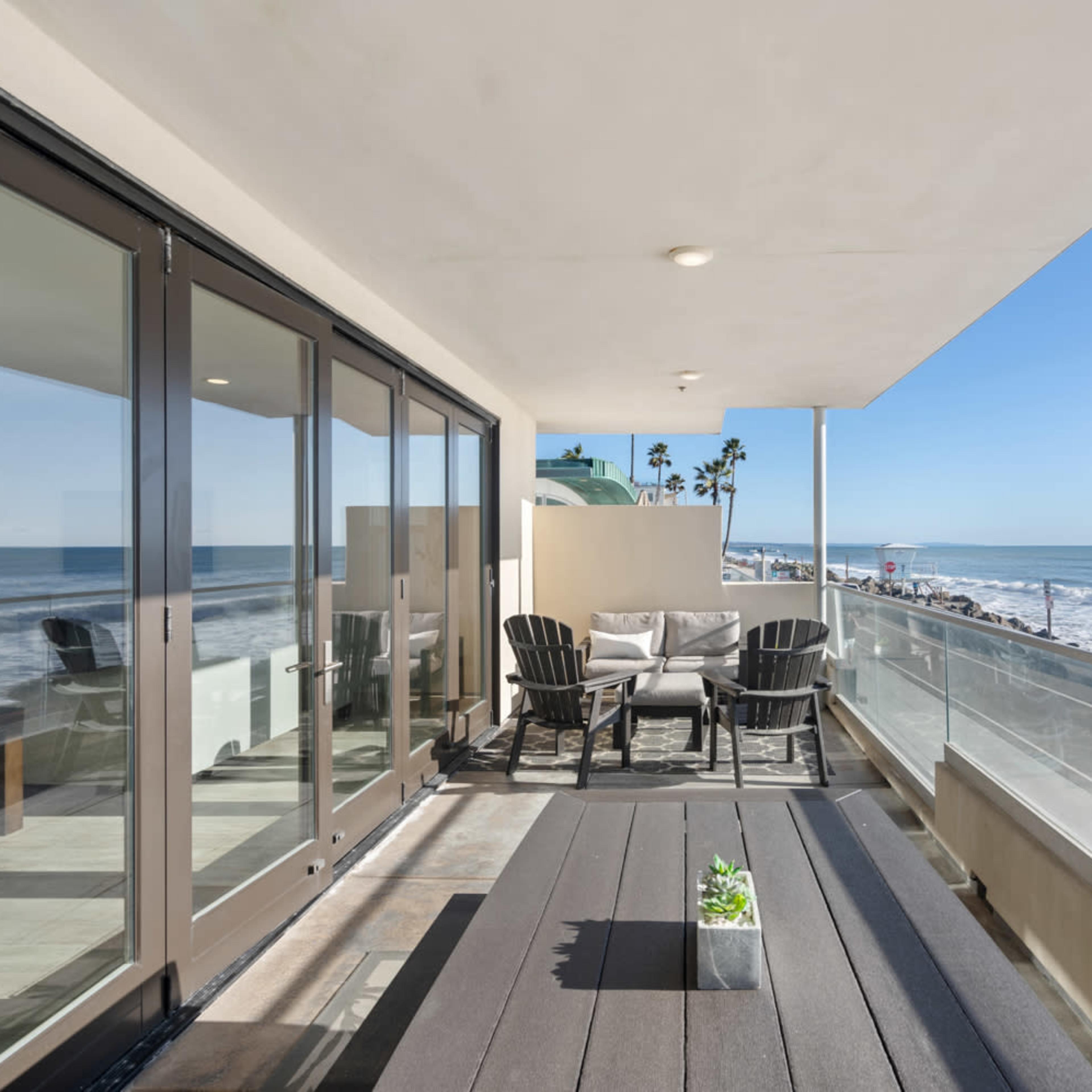 A balcony overlooking the ocean features a seating area with chairs and a table, surrounded by a glass railing with views of the beach and palm trees.