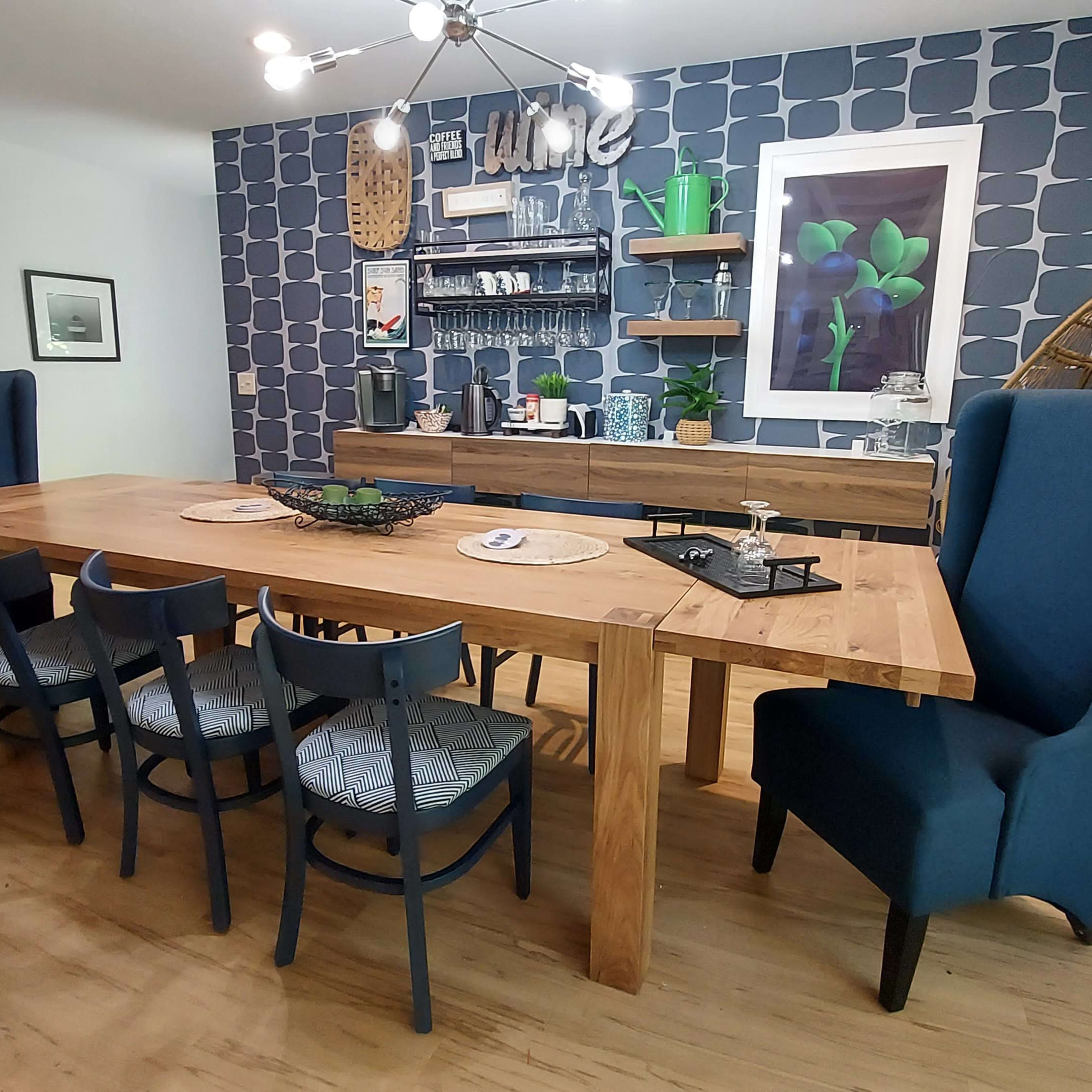 A spacious dining area with a large wooden table surrounded by high-backed chairs, set against a wall with blue patterned wallpaper and a wooden sideboard.
