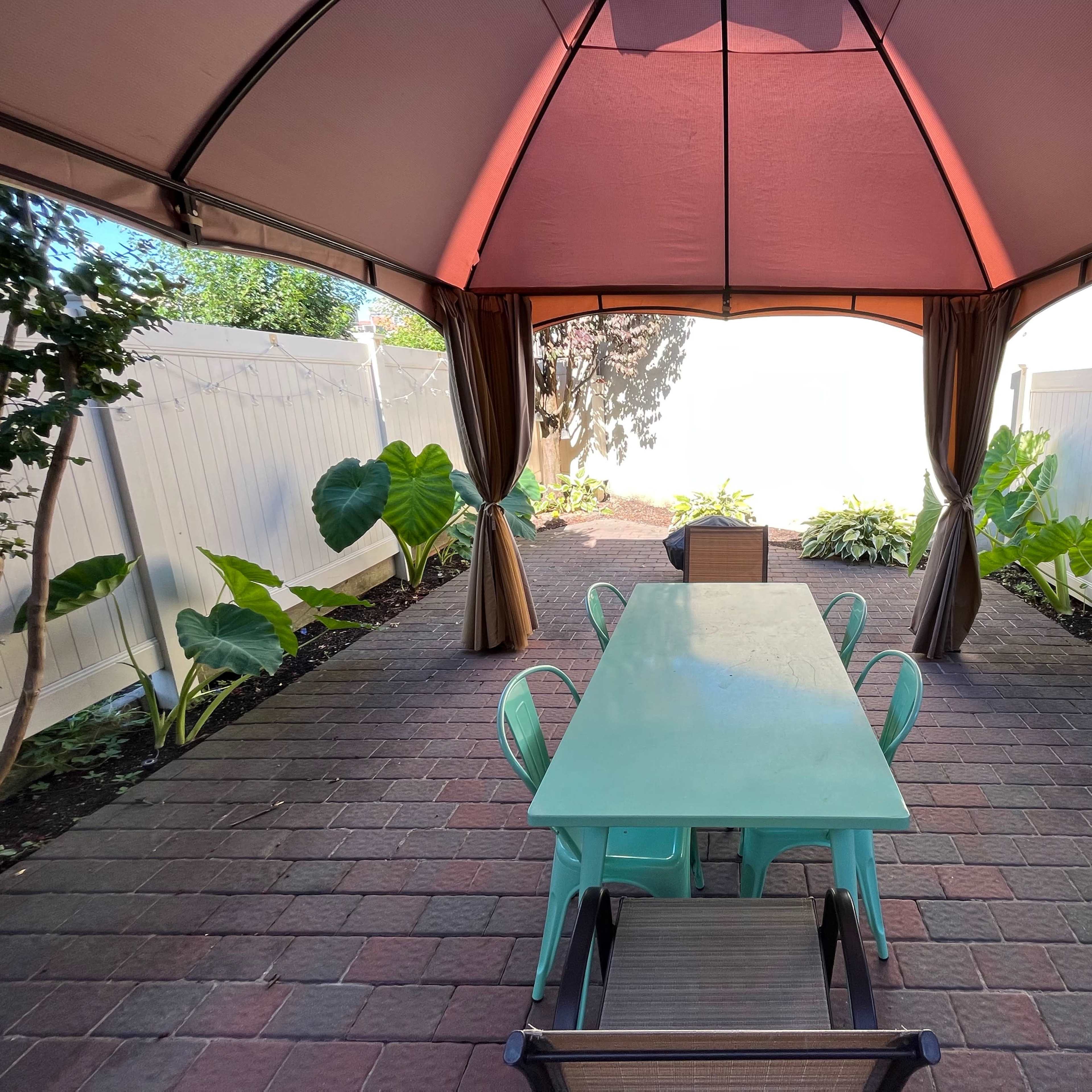 A covered outdoor seating area features a green dining table surrounded by patio chairs and bordered by large tropical plants.