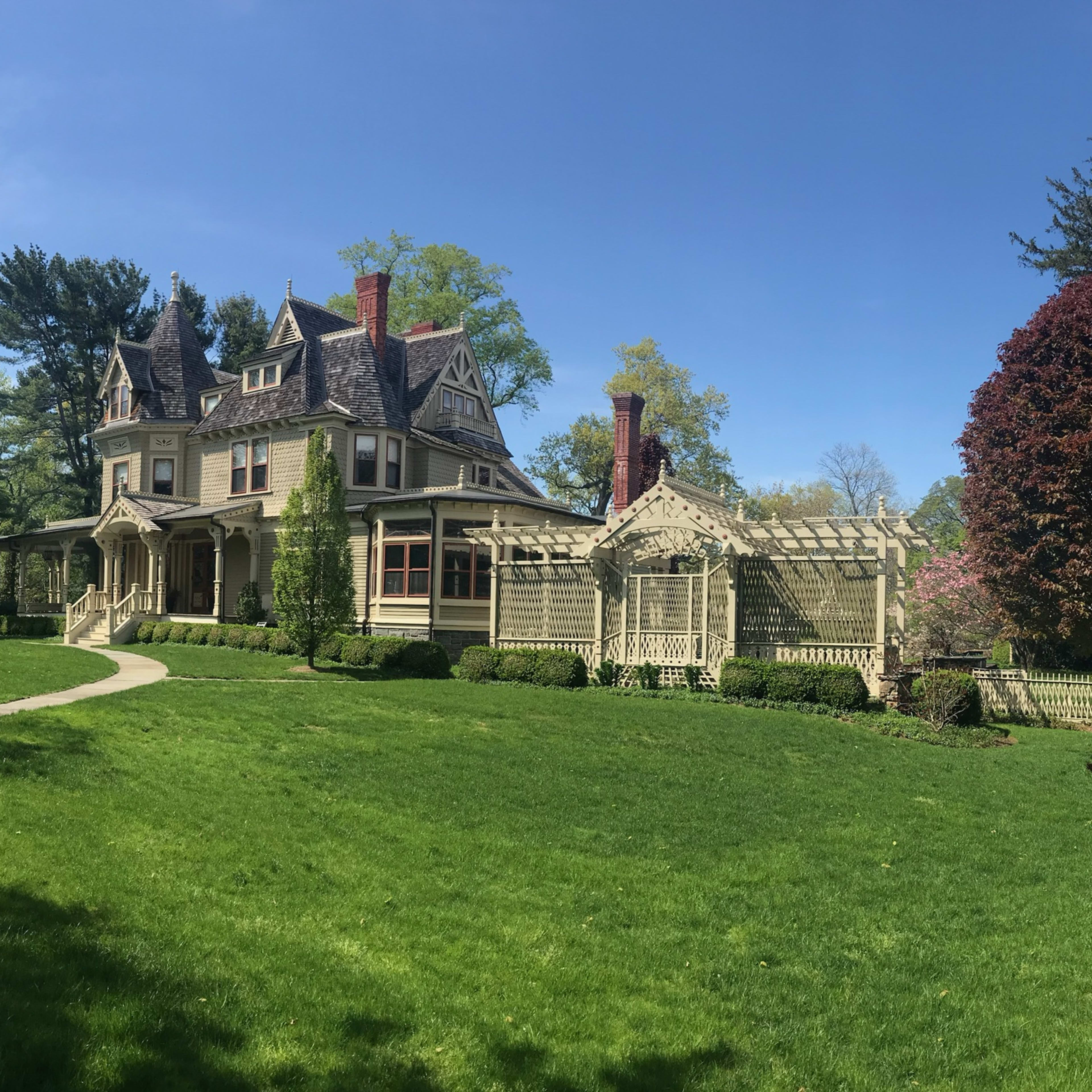 A large Victorian-style house with intricate architectural details stands beside a manicured lawn and colorful trees under a clear blue sky.
