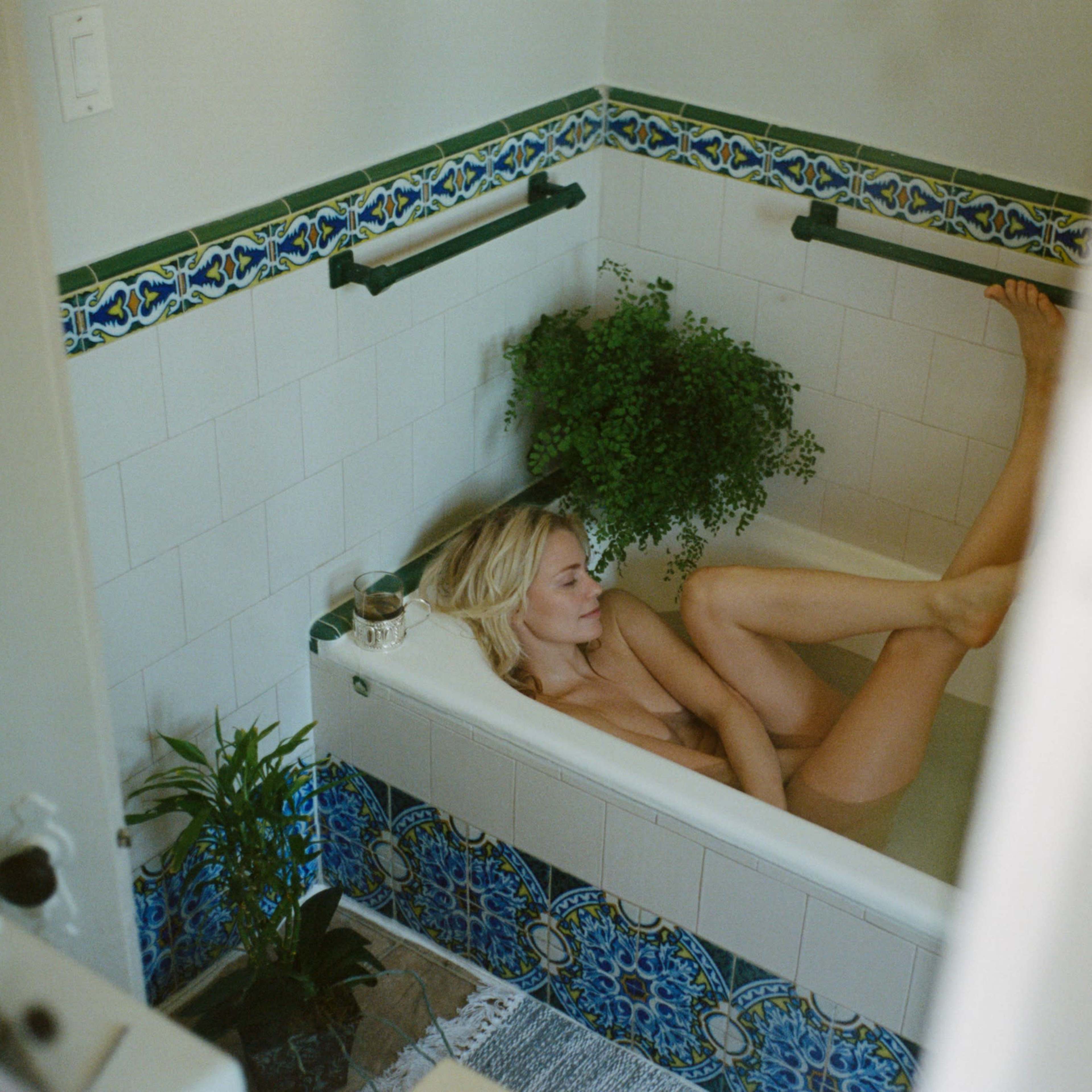 A person is sitting in a bathtub with plants around, posing with their legs drawn up.