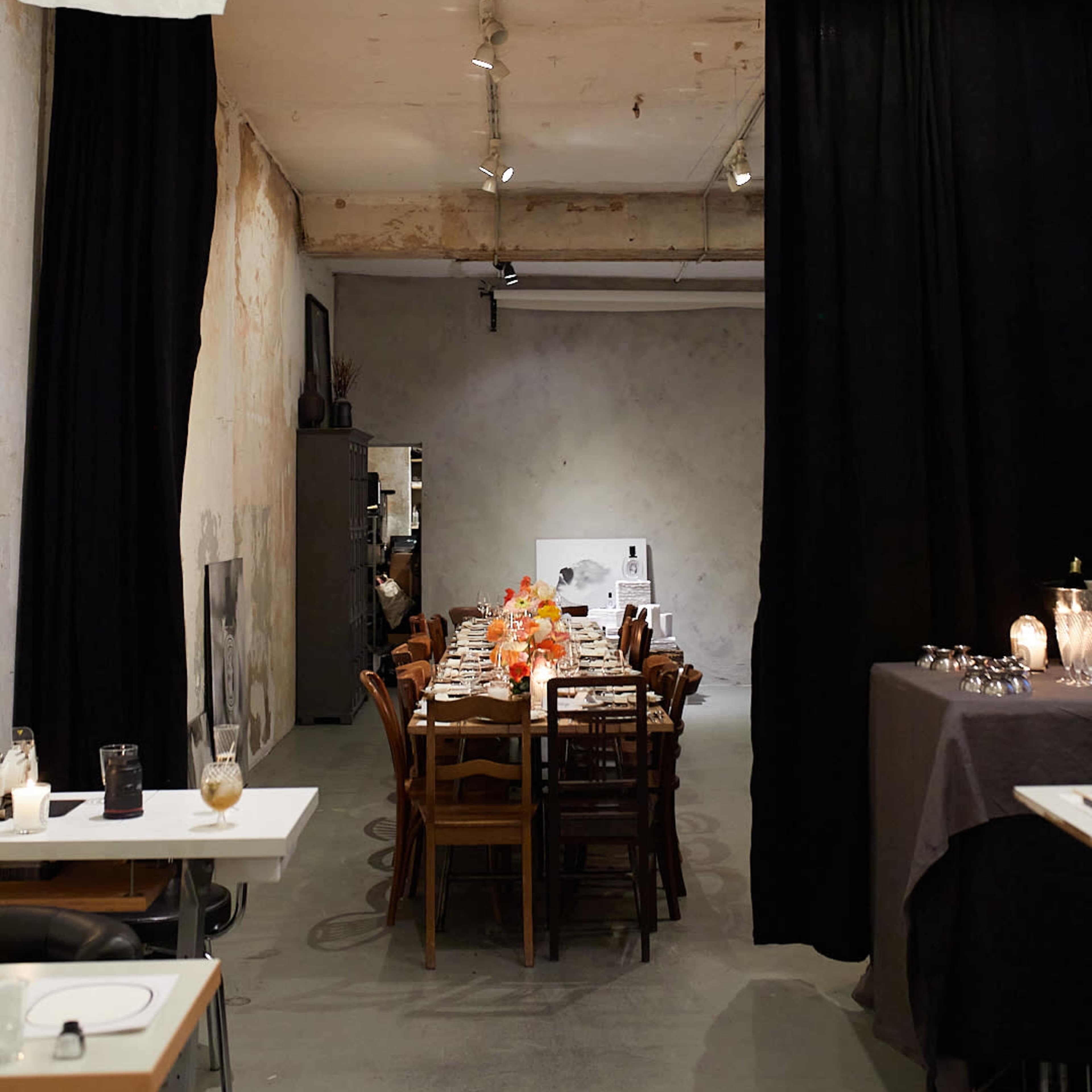 A dining area featuring a long table set with dinnerware, surrounded by wooden chairs, and framed by black curtains and a textured wall.