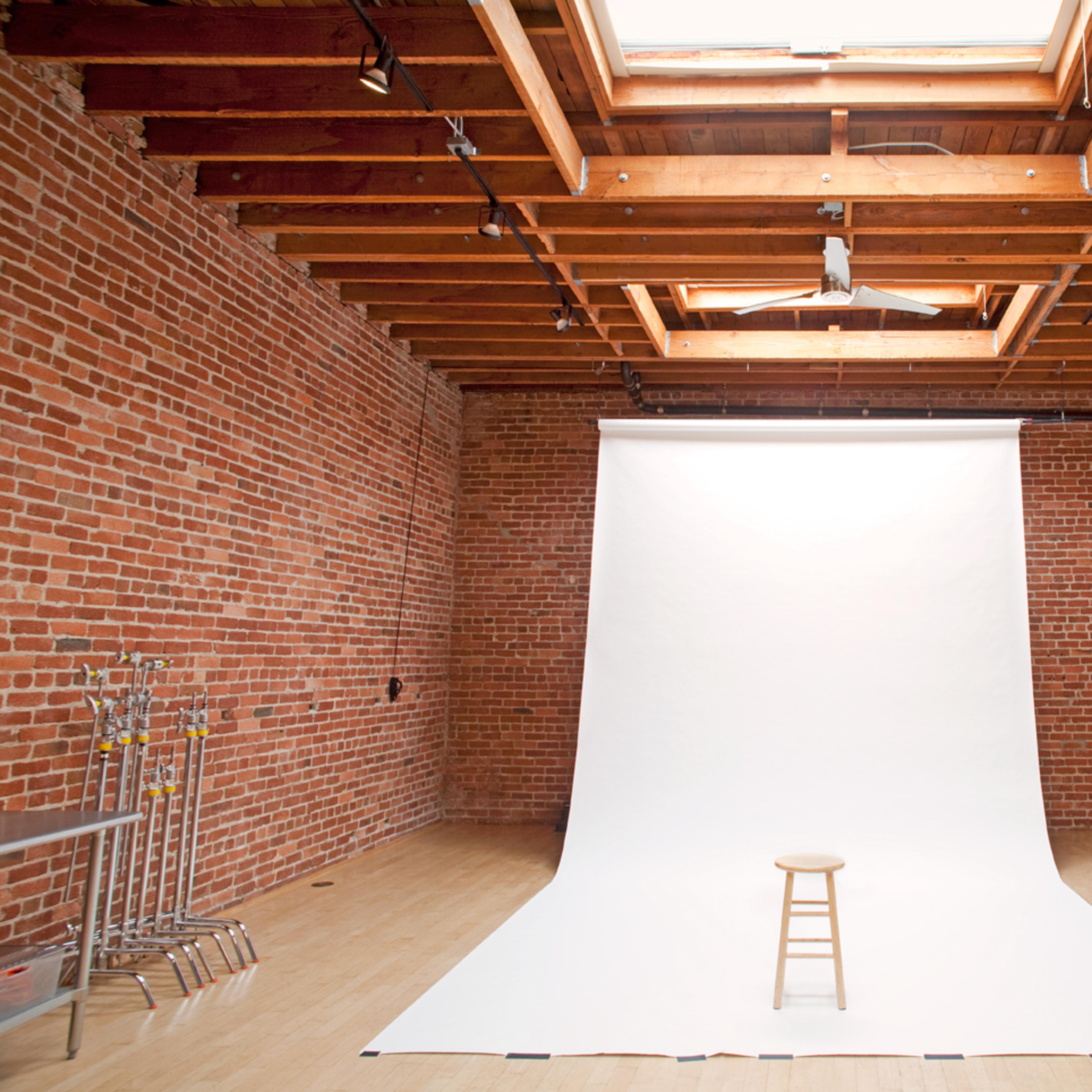 A photography studio with exposed brick walls, a white backdrop, a wooden stool, and a skylight.