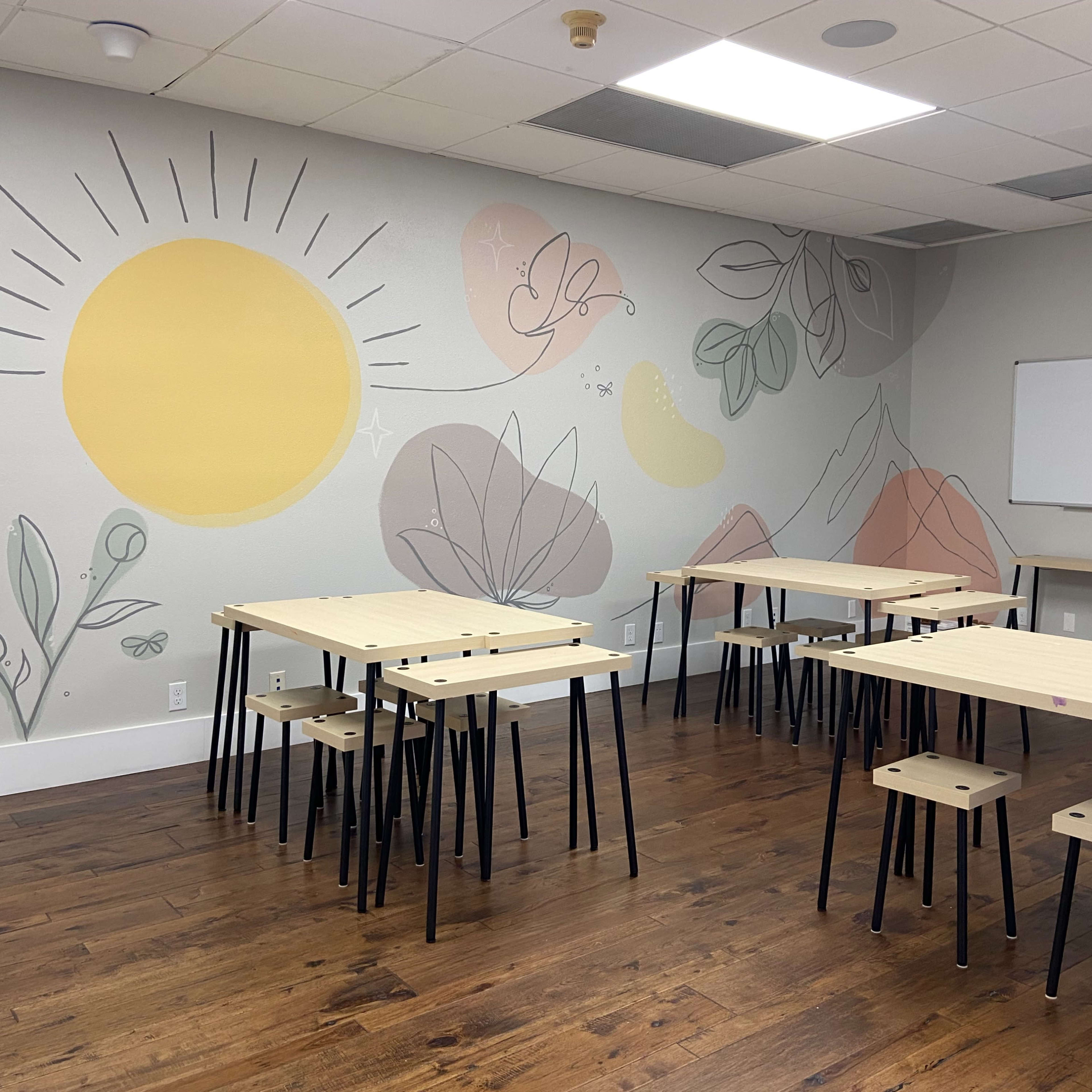 The room features light-colored walls with a mural of a sun and abstract plants, along with several wooden tables and stools arranged for seating.