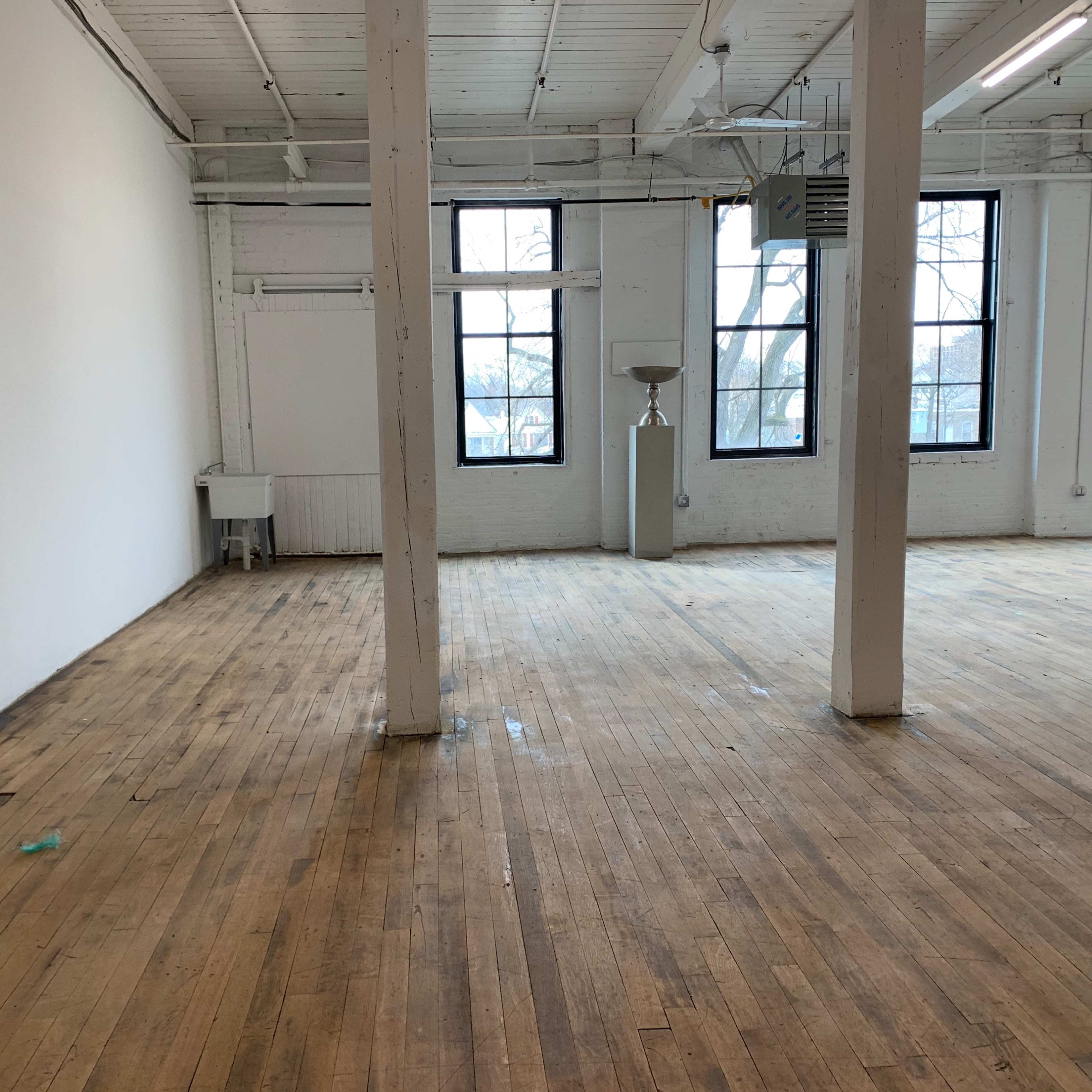 An empty room with wooden flooring, white walls, large windows, and exposed beams.
