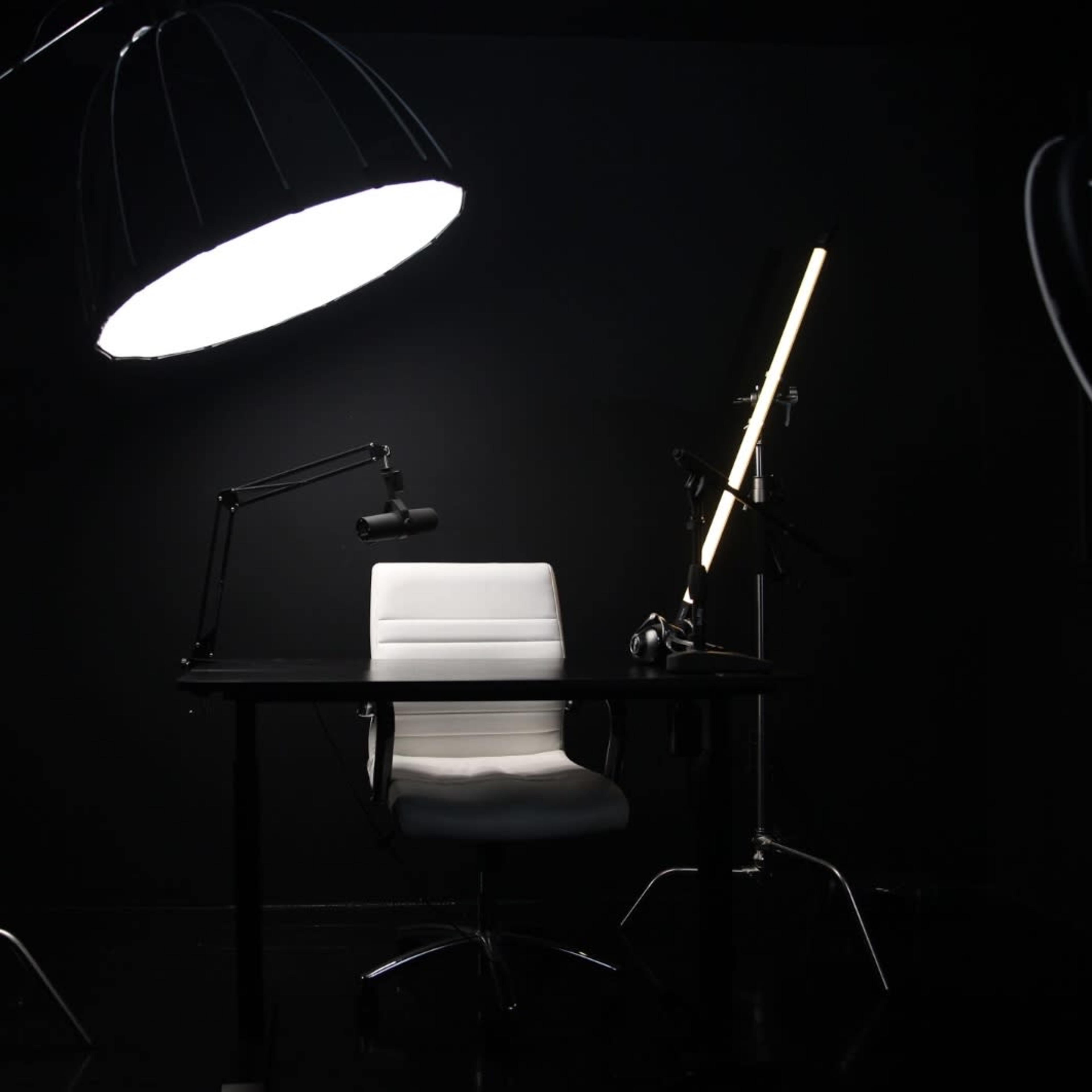 A modern office setup features a white ergonomic chair positioned at a sleek black desk, illuminated by a large overhead light and a standing floor lamp against a dark background.