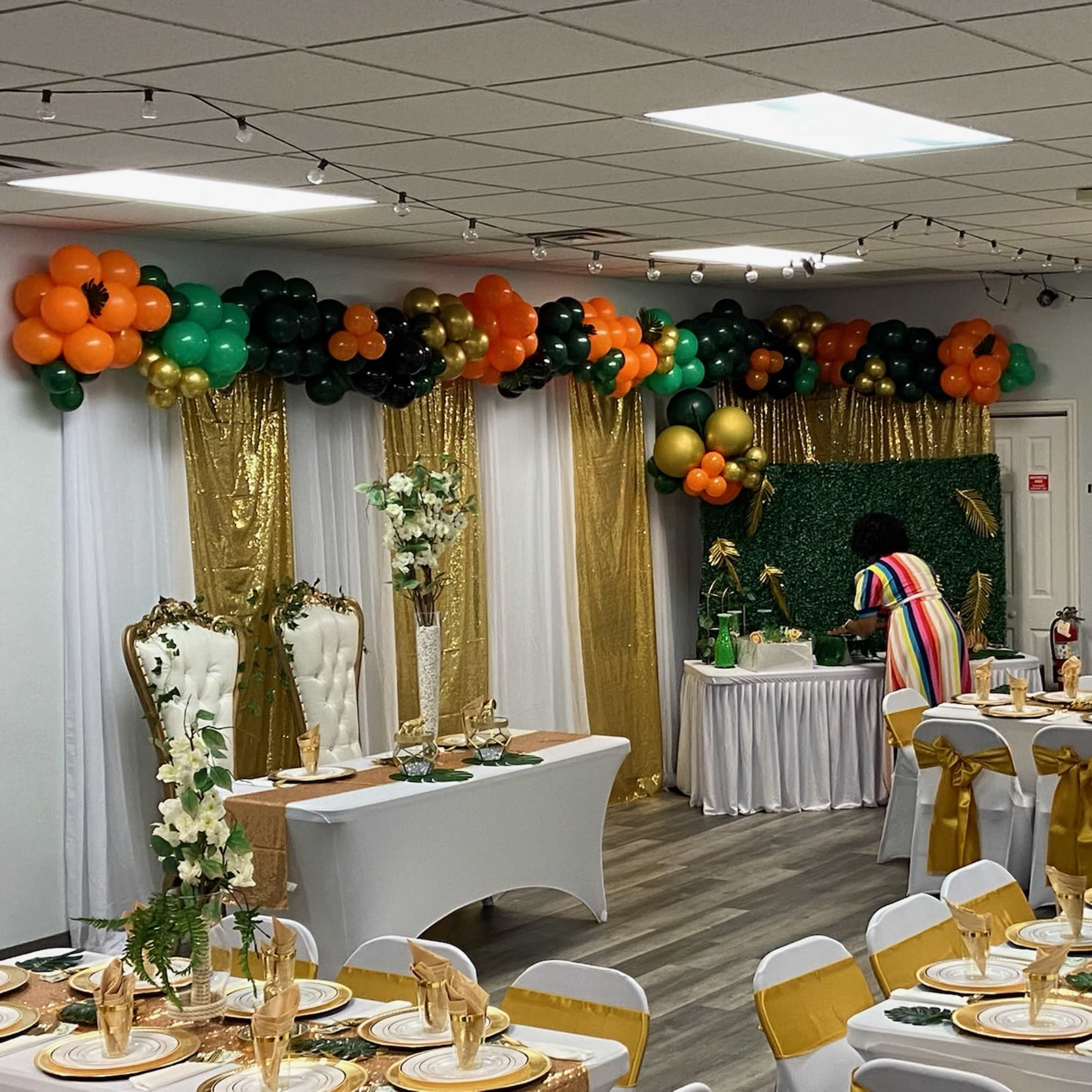 A decorated event space with tables set for a formal gathering, featuring gold and green decorations, balloons, and a person arranging items on a table in the background.
