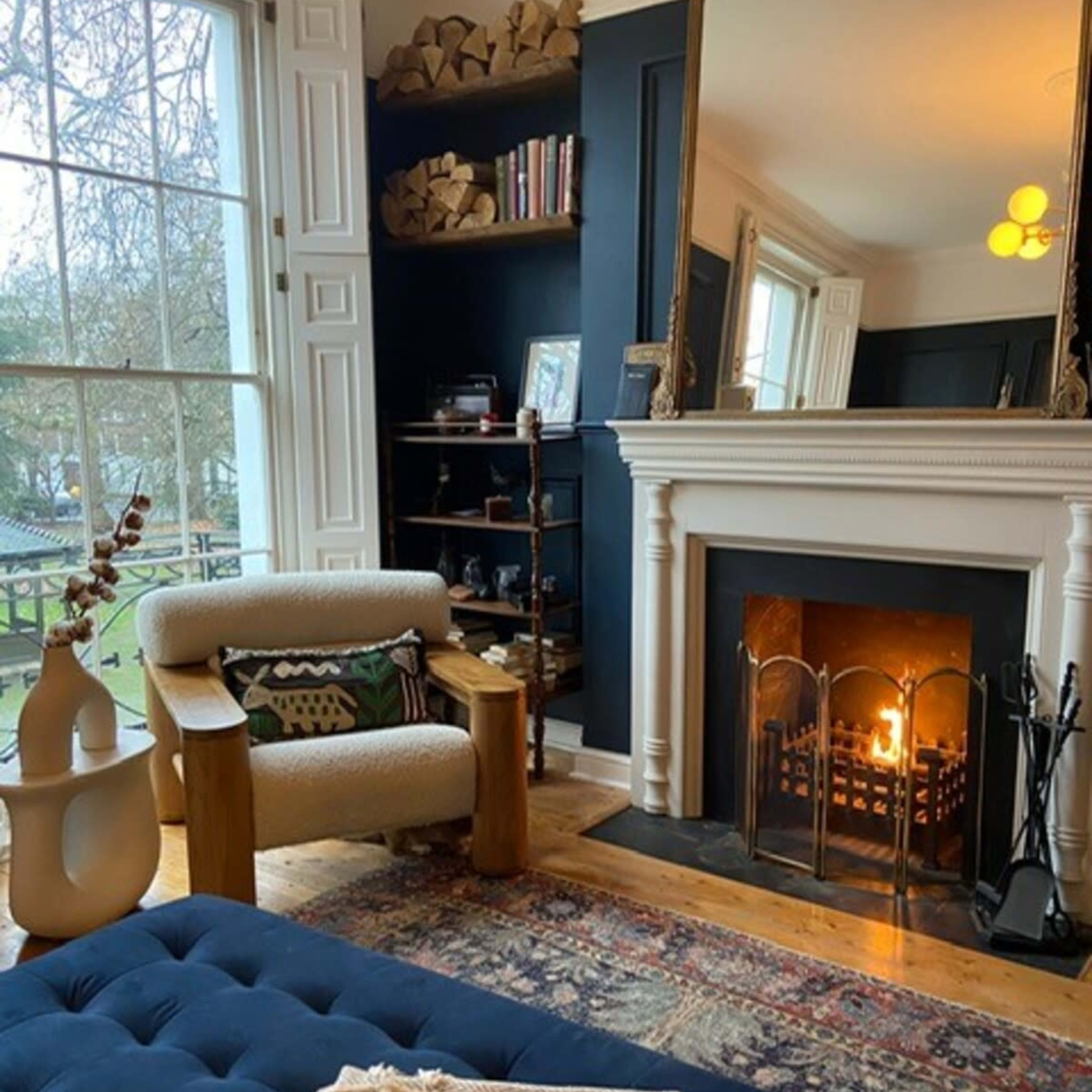 A cozy living room with a fireplace, a tufted blue ottoman, and a mirror above the mantel, overlooking a view outside through a large window.