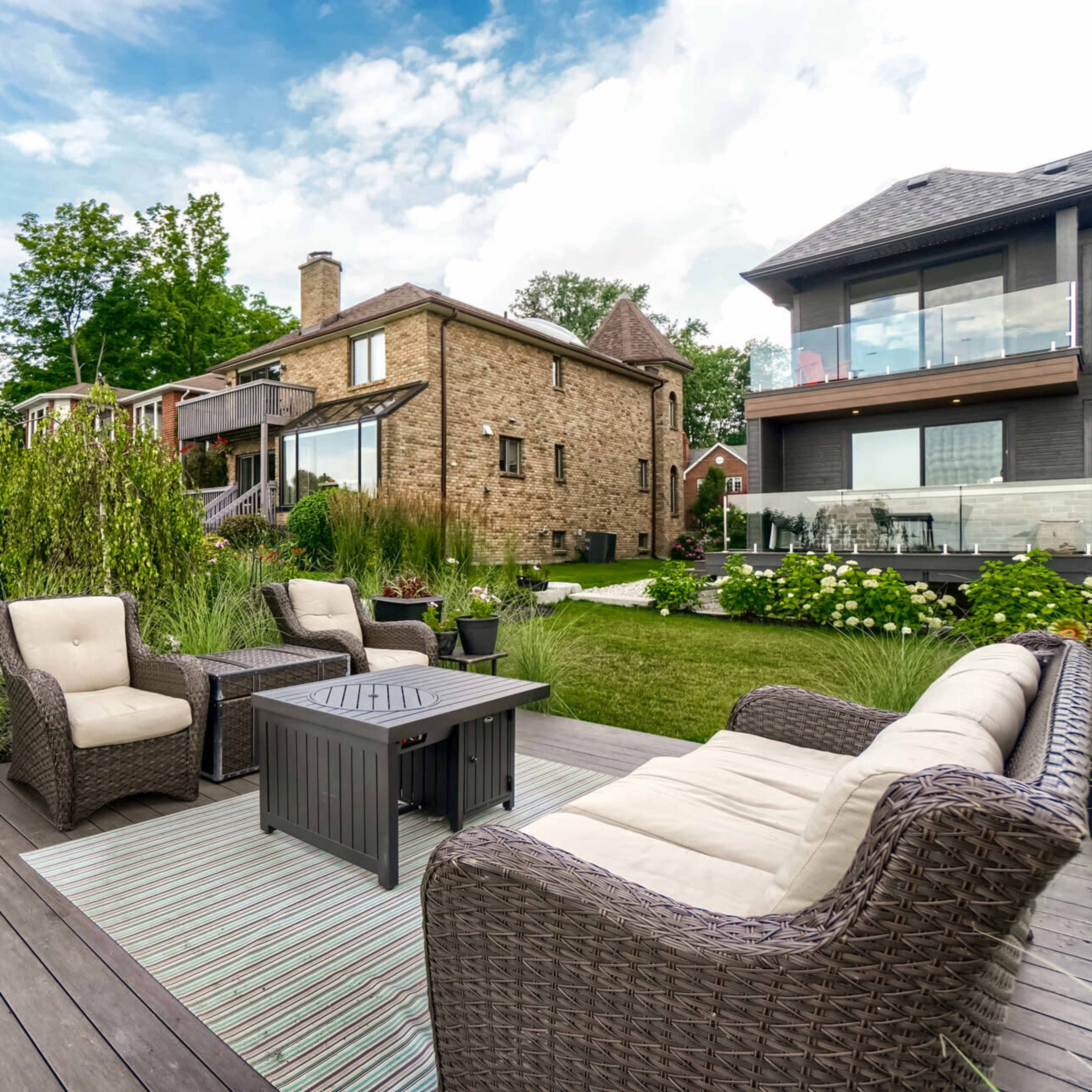 A backyard patio features a seating arrangement with wicker furniture, a coffee table, and a swing, surrounded by greenery and houses in the background.