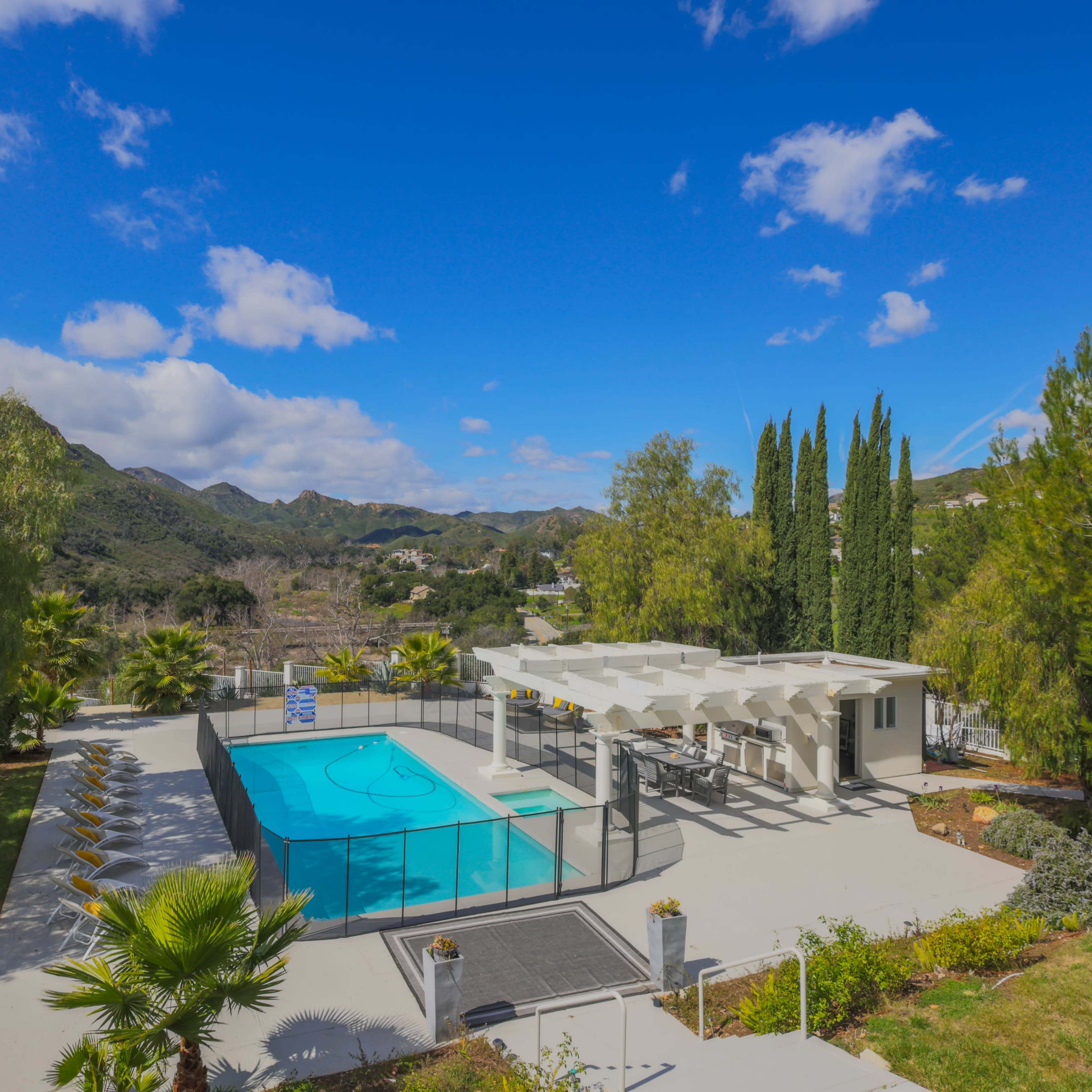 A large swimming pool surrounded by a landscaped garden and mountain views under a clear blue sky.