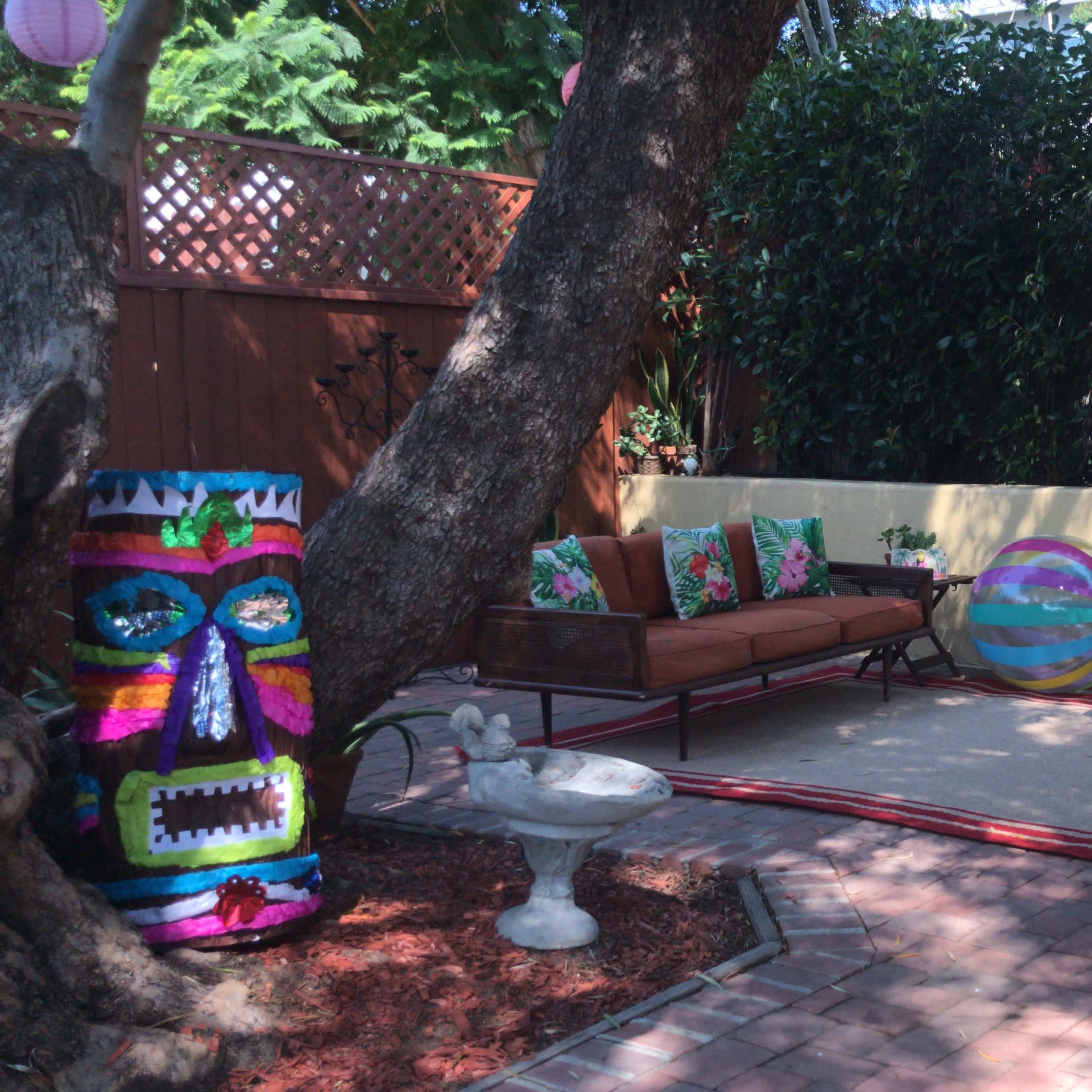 A vibrant outdoor seating area features colorful sofas and cushions, surrounded by greenery and decorative elements like a tiki statue and lanterns.