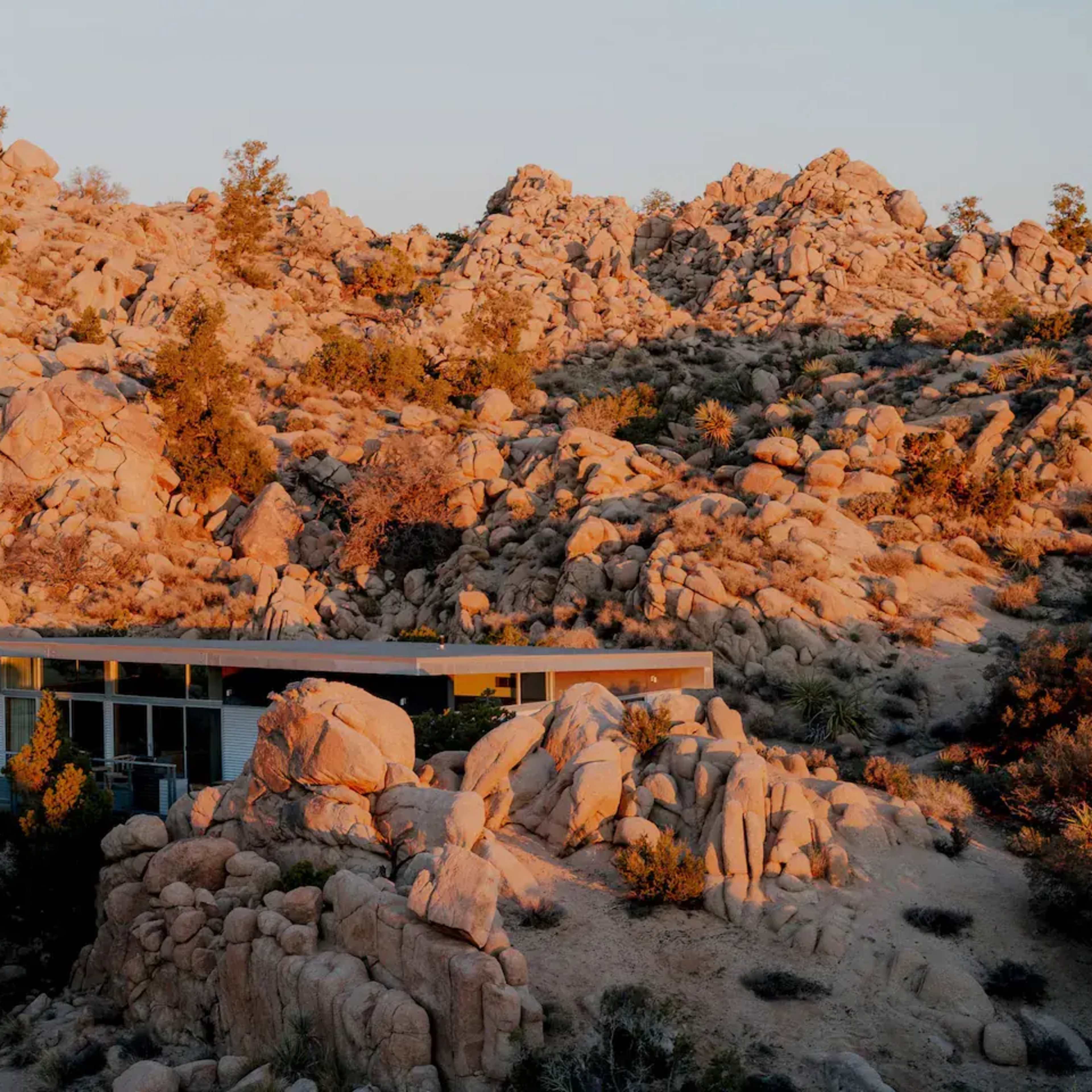 A modern house is nestled among large boulders and desert vegetation in a rocky landscape.