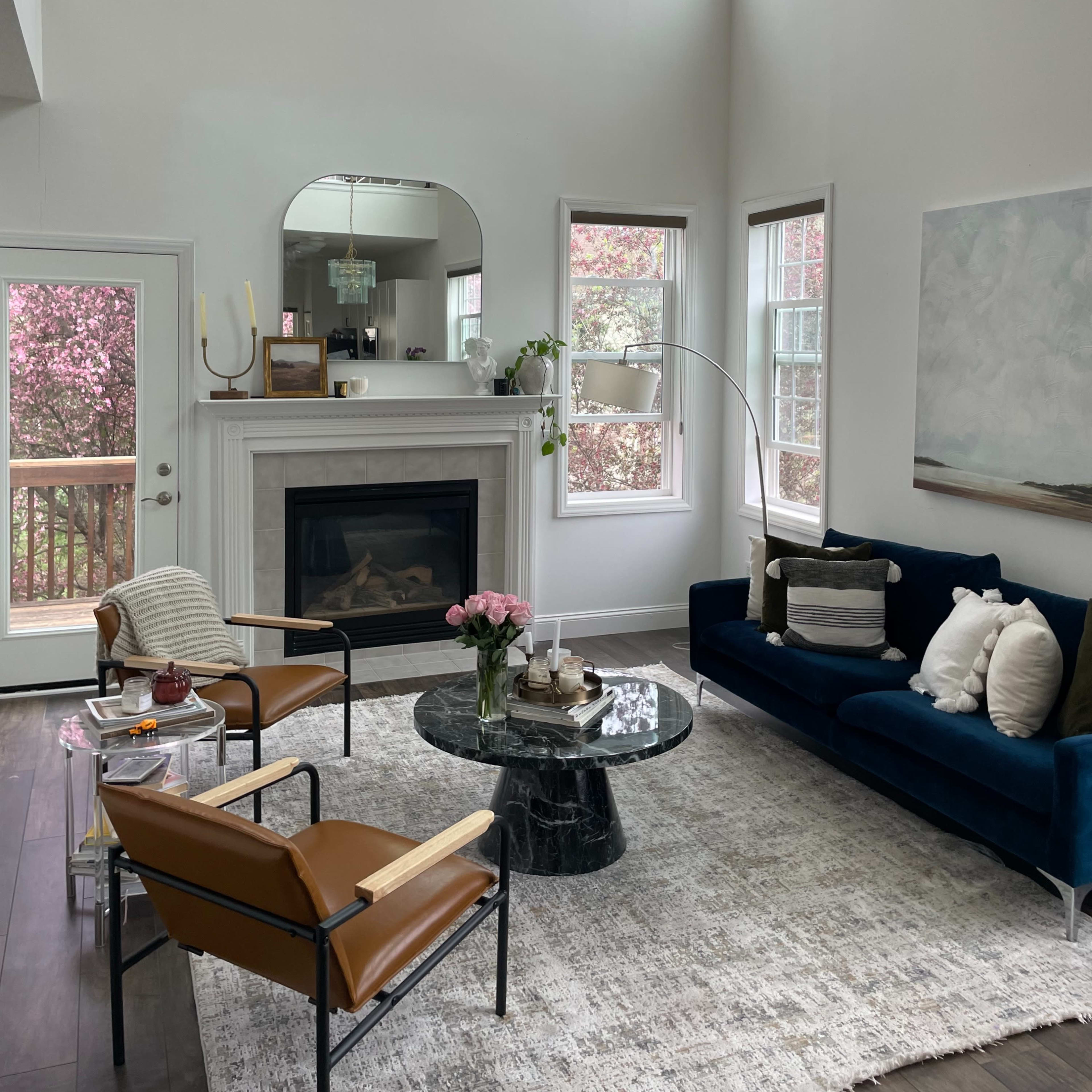 A modern living room features a navy blue sofa, two brown armchairs, a round black marble coffee table, and a fireplace, with large windows allowing natural light and a view of blooming trees outside.