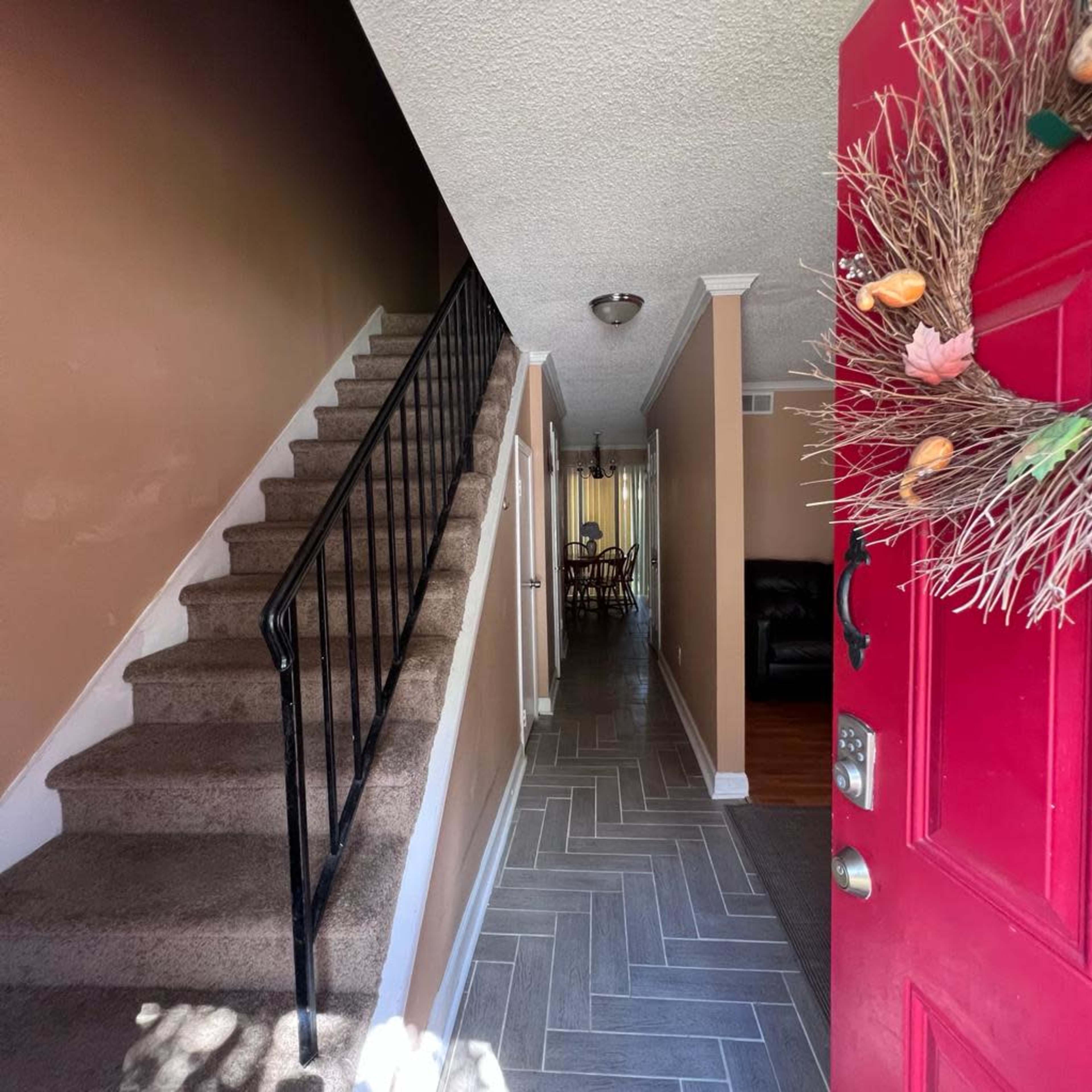 A view from an open red front door into a house, featuring a staircase on the left and a hallway leading to a dining area in the background.