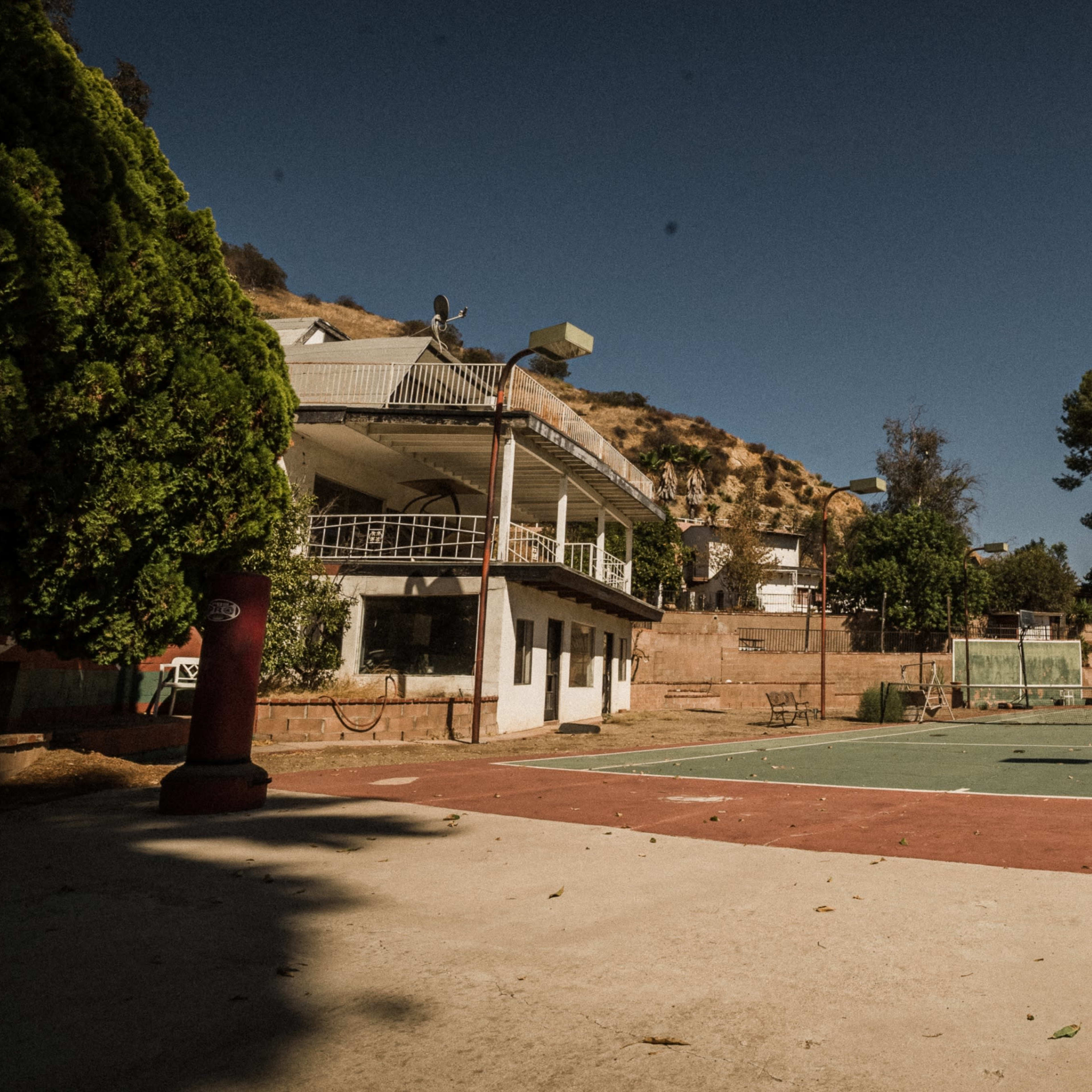 A vacant tennis court surrounded by a two-story building and trees, set against a hilly background under a clear sky.