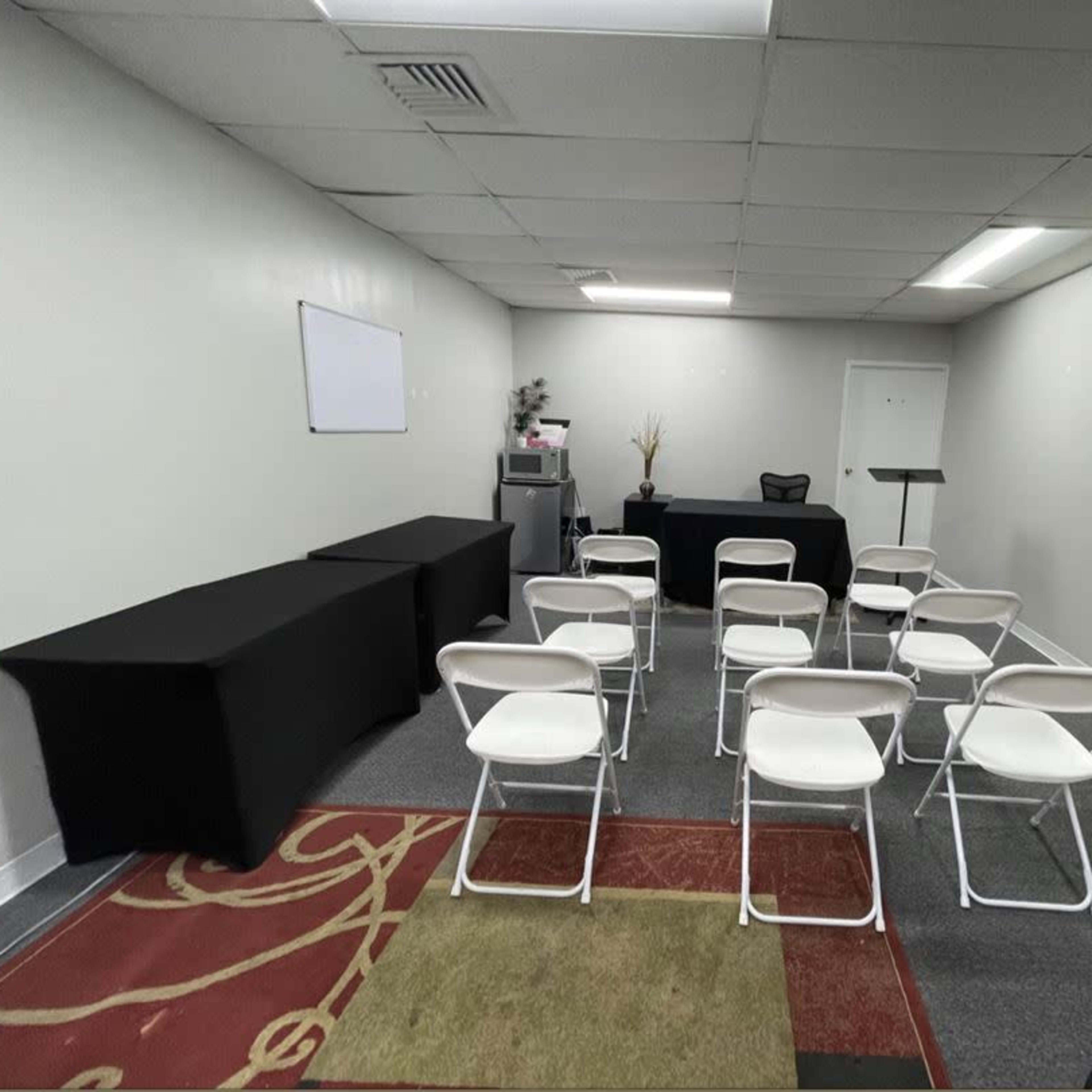 A small meeting room features several white chairs arranged in rows facing a black table and a podium against the wall.