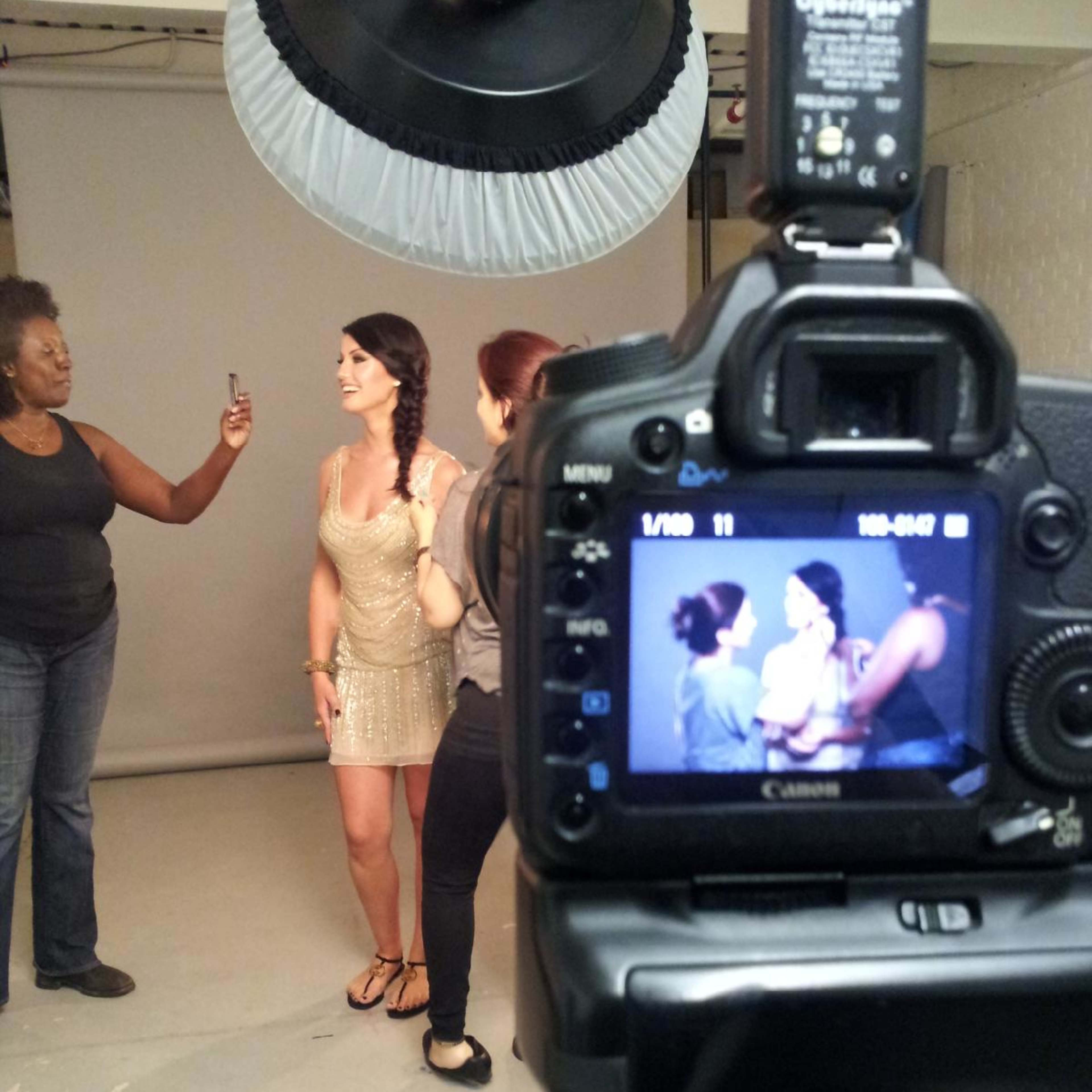 A camera captures a photoshoot scene where a model in a dress is posing while being assisted by two stylists in a studio.