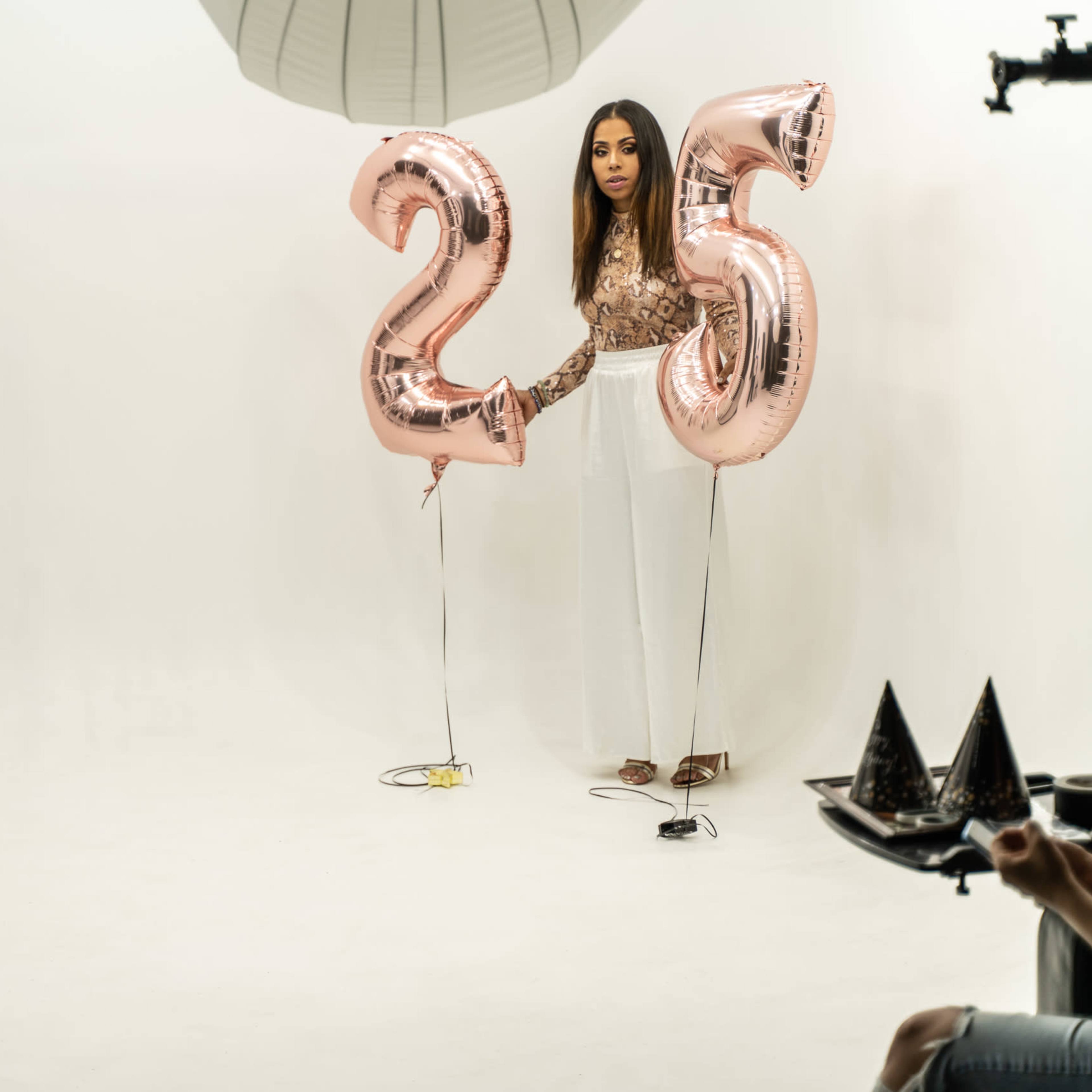 A man stands on one side of a studio while a woman holds two large rose gold balloons that read "25" on the other side.