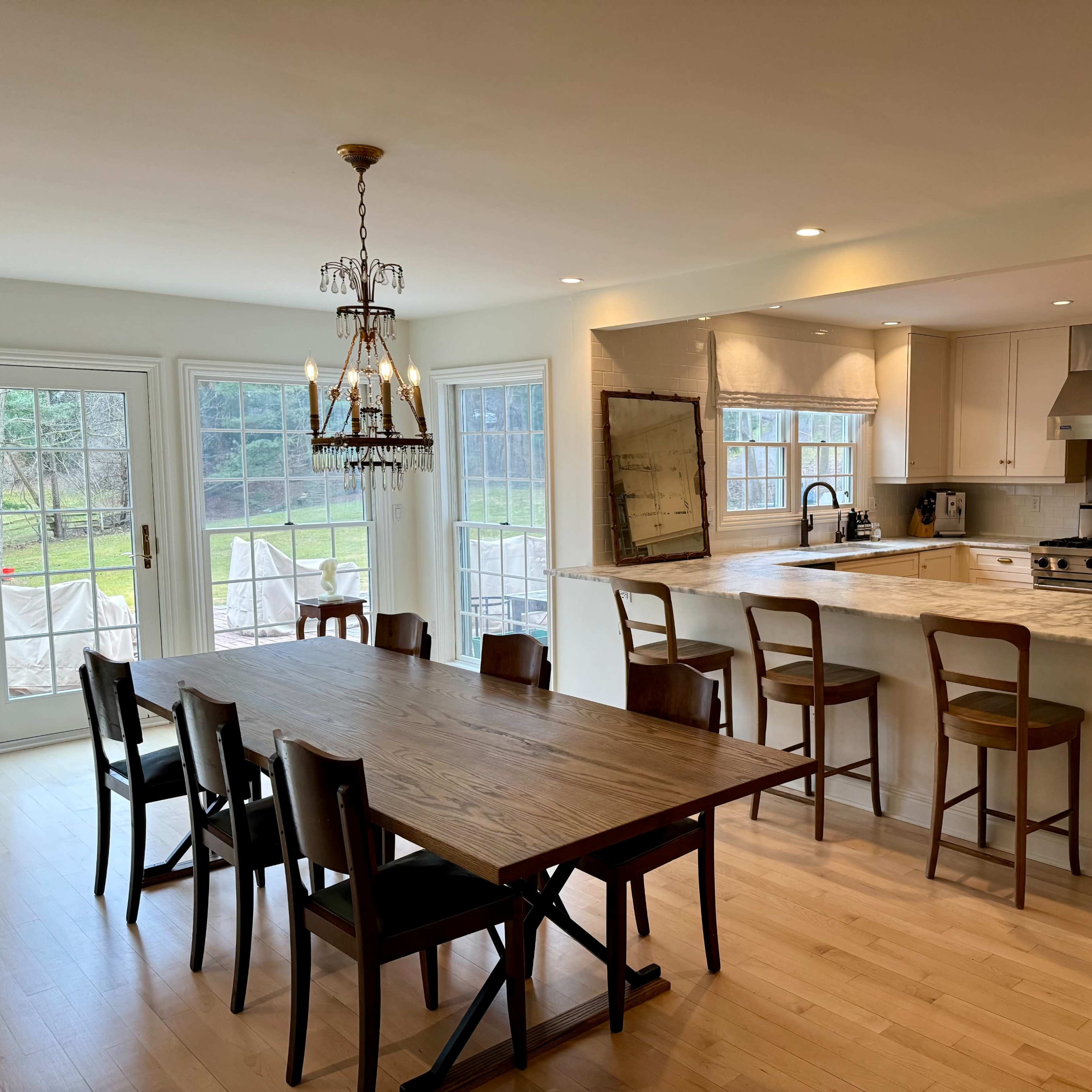 A spacious kitchen and dining area features a large wooden table with several chairs, an island with bar stools, and large windows providing a view of the outdoors.