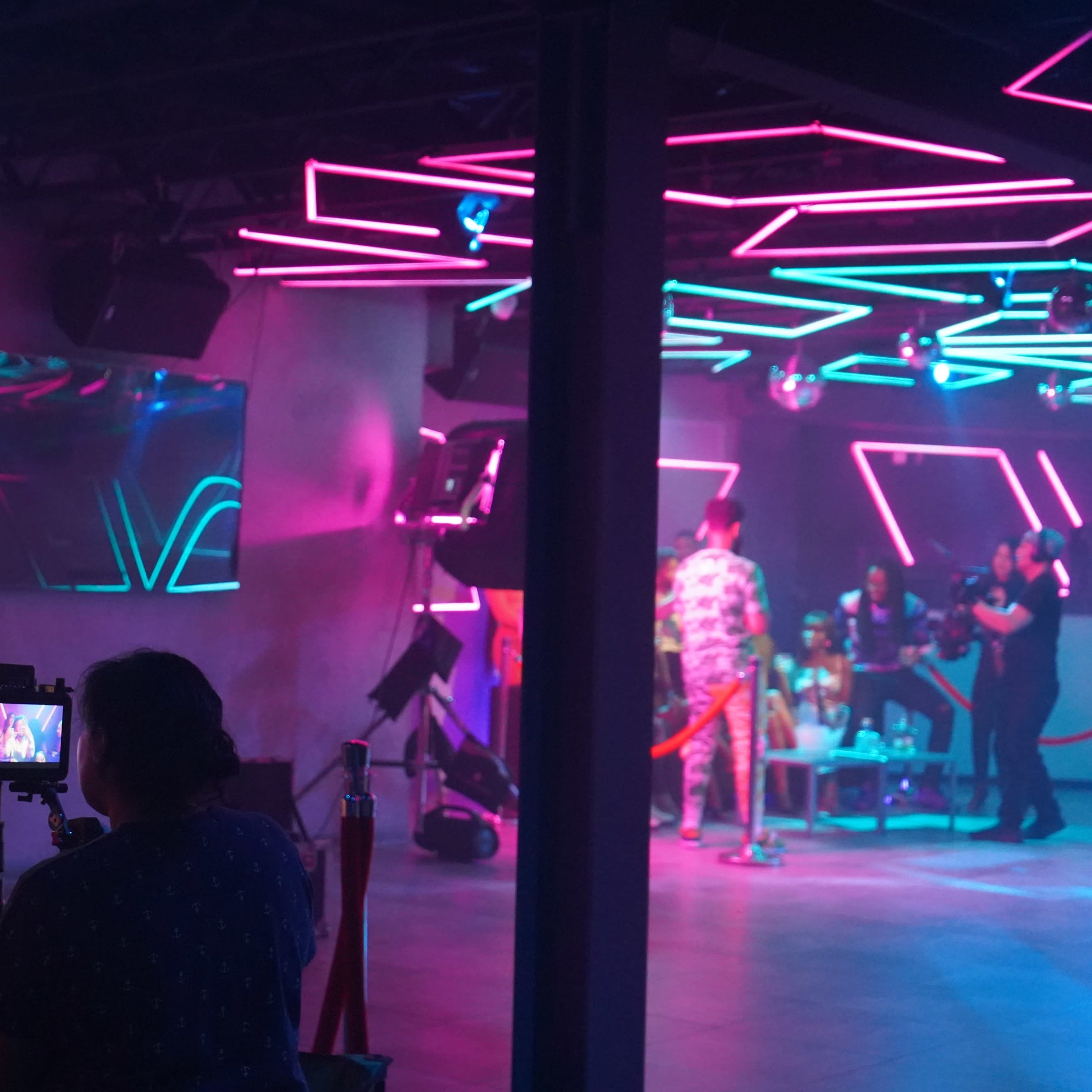 A film crew captures a colorful scene in a nightclub setting with neon lighting and multiple cameras positioned on tripods.