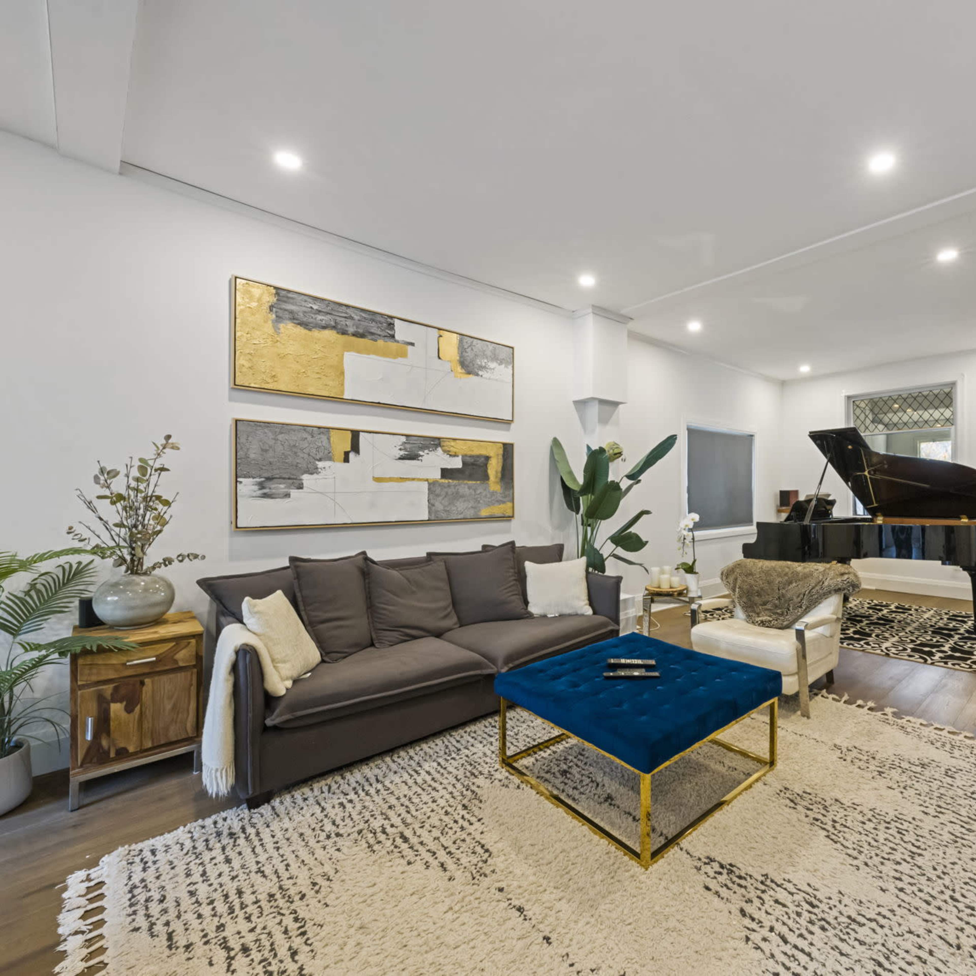 A modern living room features a black piano, a blue coffee table, a large rug, and abstract wall art against white walls.