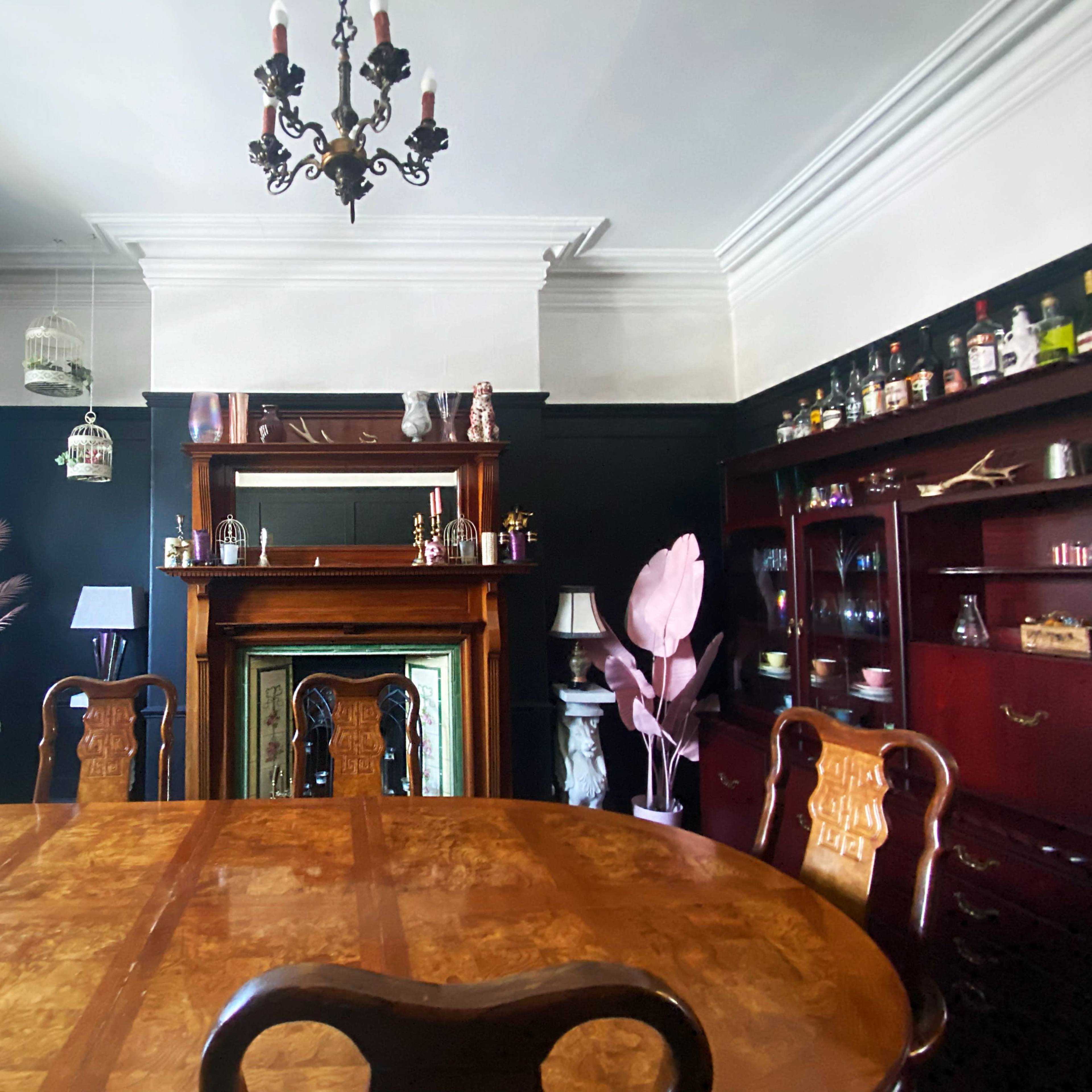 A wooden dining table is positioned in a room with dark walls, featuring a fireplace, a mirror, and various decorative items.