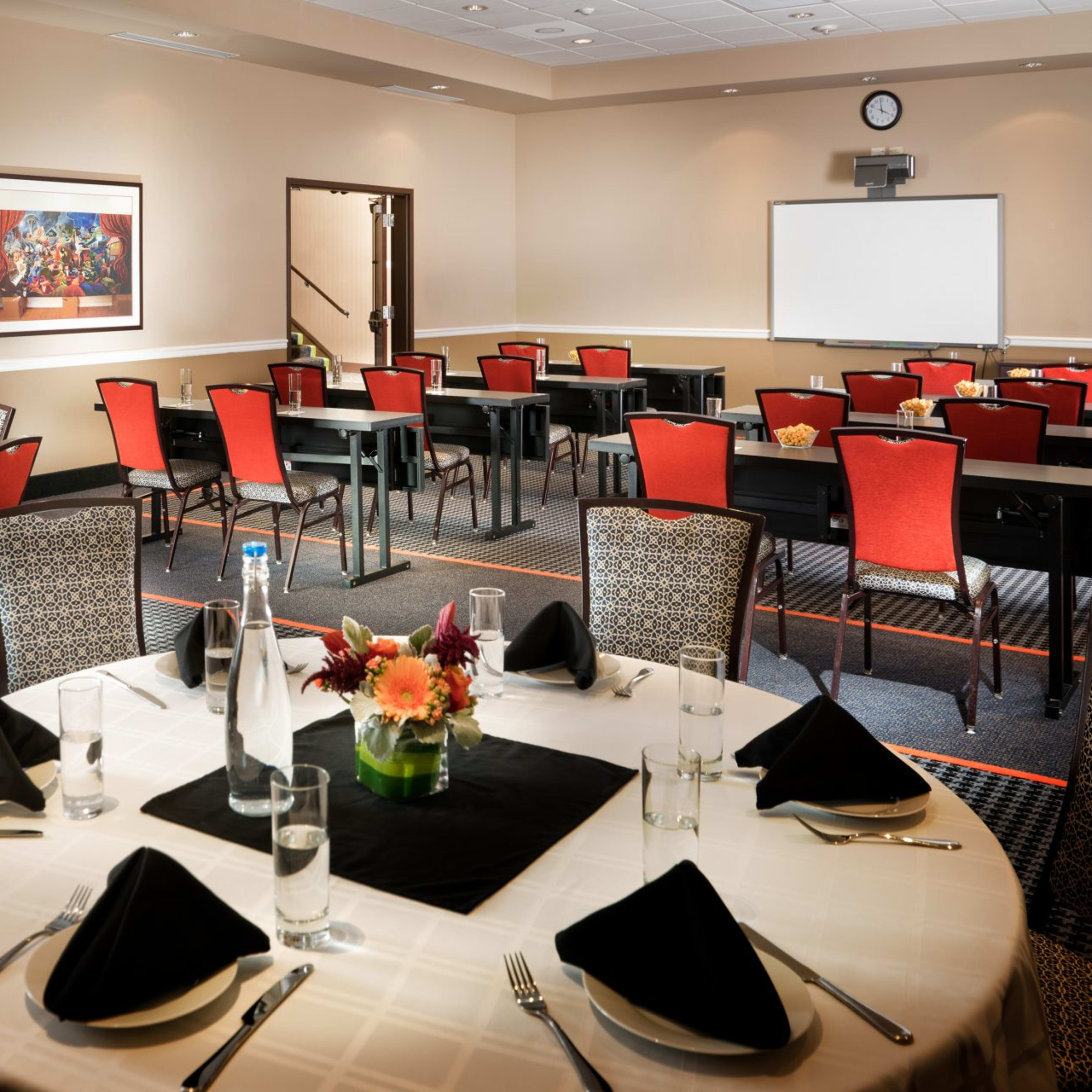 A dining area set with round tables and chairs in a well-lit room, featuring floral centerpieces and a presentation screen at the front.